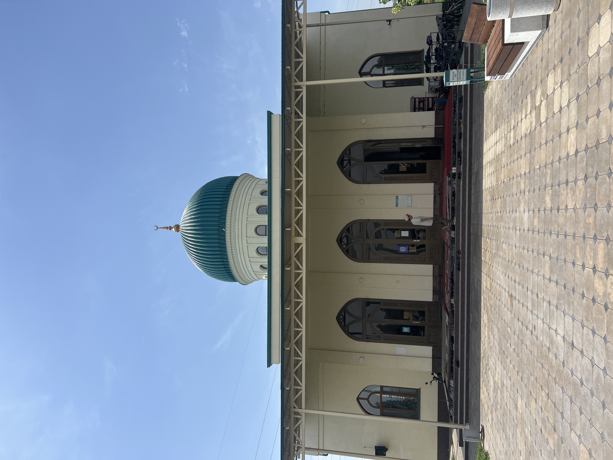 In the city of Karamanas, Uzbekistan, a mosque with a prominent green dome stands under a clear blue sky. The mosque features a cream-colored facade with arched windows and doors. A covered walkway extends from the building, supported by white metal beams. In front of the mosque, a paved courtyard leads to steps where prayer rugs are laid out, and numerous pairs of shoes are neatly arranged, indicating recent or ongoing prayer services. A lone individual wearing a white cap and a light-colored shirt walks on the steps. To the right of the entrance, a bench and a trash receptacle are visible. The overall scene conveys a sense of peace and religious devotion.