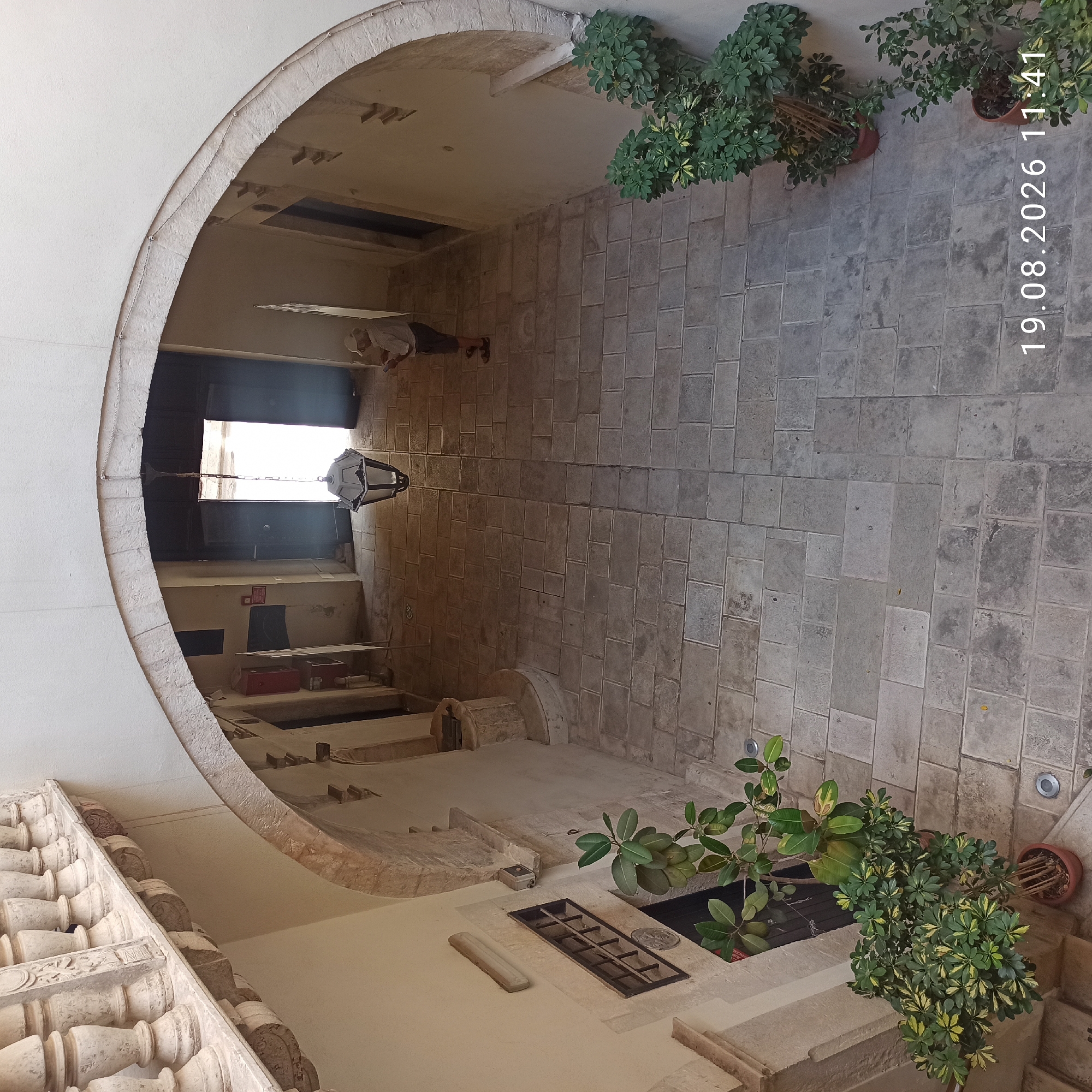 The image shows an interior courtyard with arched doorways and stone paving. A person in a light-colored shirt and shorts is standing near an open doorway, looking at their phone. There are potted plants on either side of the courtyard. A vintage street lamp hangs from the ceiling. The architectural style suggests a historical building. The date stamp "19.08.2026 11:41" is visible in the lower right corner. This scene is likely located in the historic center of Ruvo di Puglia, Italy, given the architectural elements and the regional style.