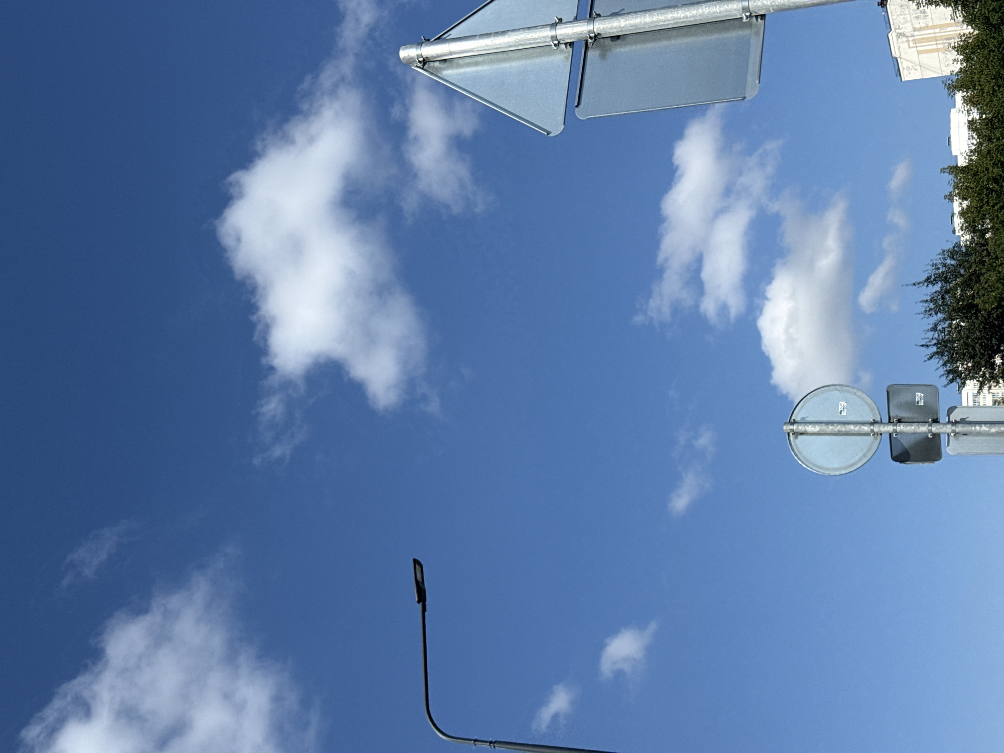 The image captures a bright, sunny day in Lyubertsy, Russia. The sky is a vibrant blue, dotted with fluffy white clouds. In the foreground, a modern street lamp with a sleek design is positioned on the left side of the frame. To the right, a metal pole supports two road signs, one triangular and one circular. Below these signs, a cluster of lush green trees partially obscures buildings in the background. The overall scene suggests an urban environment with clear weather conditions.