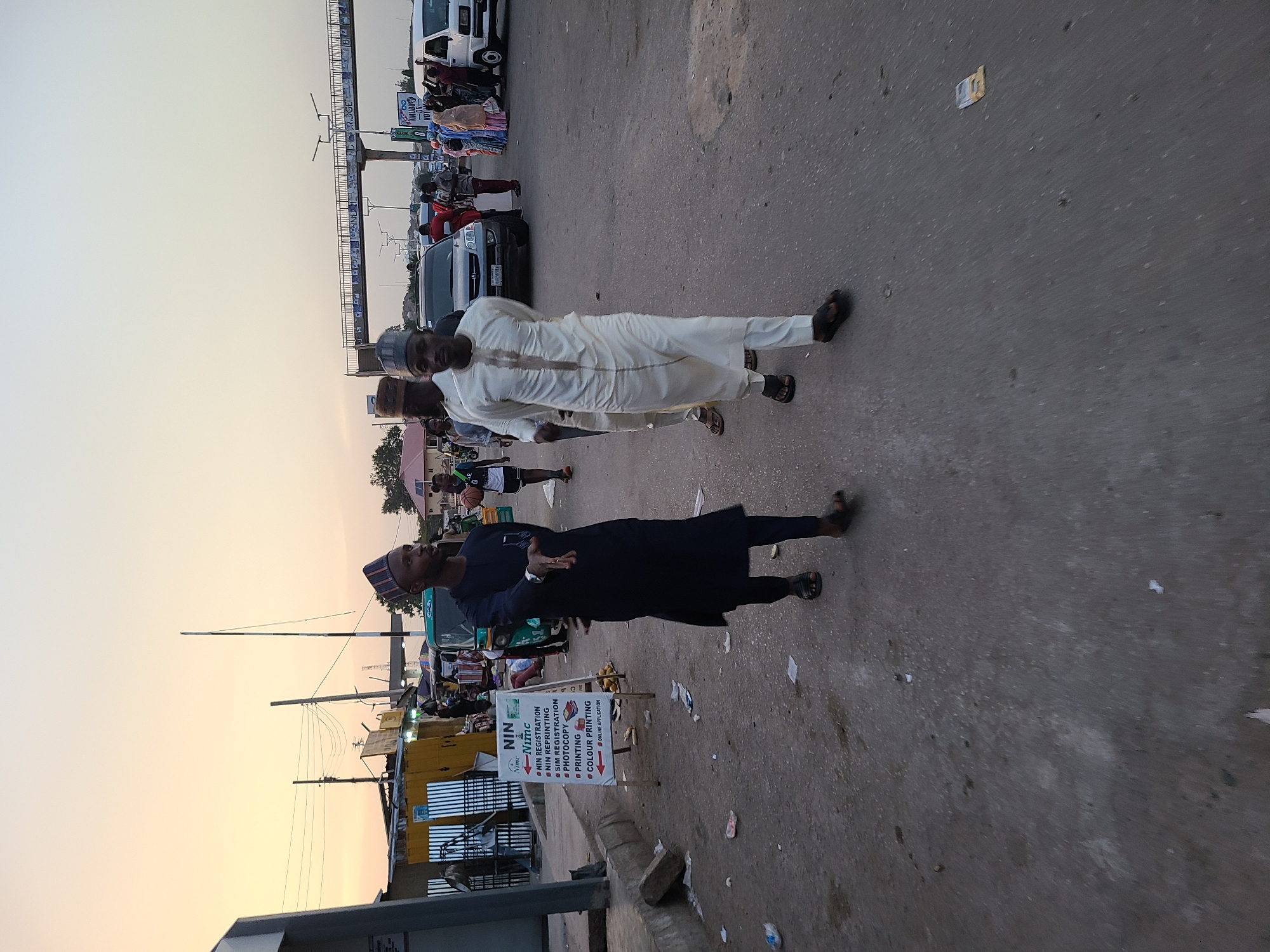 The image depicts a street scene in what appears to be a city in an unknown location. Three men wearing traditional white attire and dark sandals are walking on the right side of the frame. Another man, wearing a dark blue outfit and a striped cap, stands near a sign indicating services such as NIN registration, SIM registration, photocopying, printing, and color printing are available. This suggests the presence of a business or service center nearby.
In the background, there's a street filled with vehicles, including cars and vans, as well as other pedestrians. A tall structure with multiple overhead bars is visible, possibly indicating a marketplace or transportation hub. The lighting and sky suggest it's either early morning or late afternoon. The ground is asphalt with some litter.