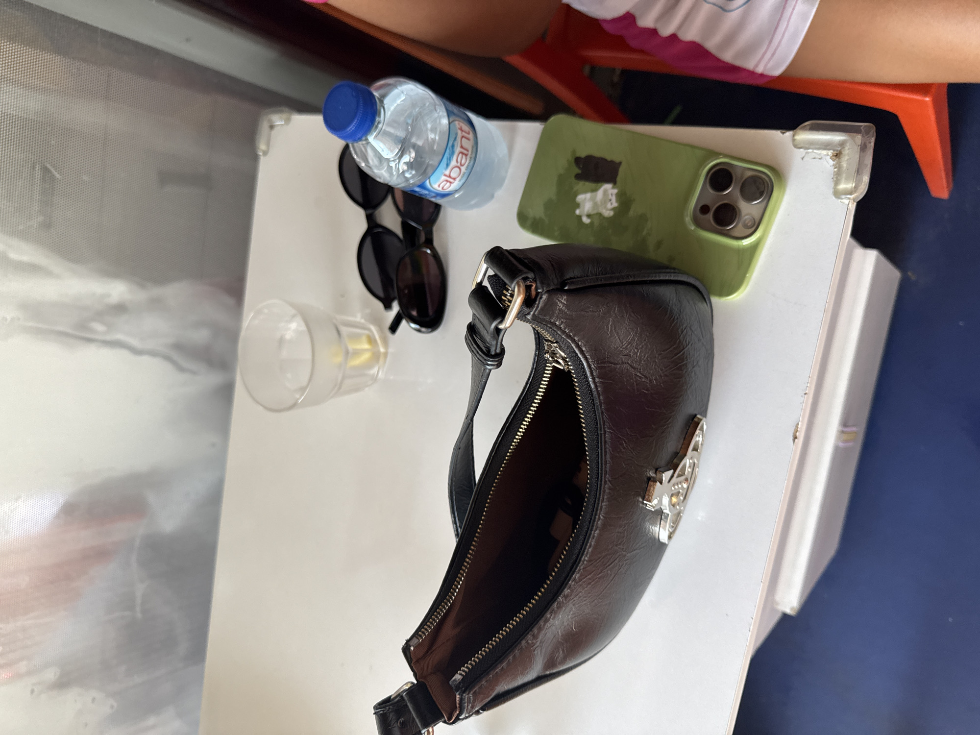 A black leather shoulder bag with a silver orb logo sits open on a white surface. Next to it are a pair of black sunglasses, a glass of clear liquid with ice and lemon, and a bottle of "abant" brand water. A green iPhone case with two silhouetted figures is also present. The scene appears to be on a table, possibly outdoors, with a dark blue surface below. The background shows a blurred reflection of what seems to be a person in a boat. The location is the city of Aksu, Turkey.