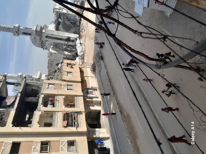 The image depicts a street scene in Khan Yunis, Palestinian Territory, characterized by significant structural damage to surrounding buildings. In the left foreground, a multi-story, light brown apartment building shows extensive damage, particularly on its upper floors, with collapsed sections exposing interior rooms. Laundry hangs on a second-floor balcony. In the right background, a large, white mosque featuring a prominent dome and two minarets is severely damaged, with large portions of its structure, including the dome and part of a minaret, appearing collapsed into rubble.
A paved road with white lane markings runs through the center of the image. Numerous individuals, including adults and children, are walking along the road and sidewalks. On the left sidewalk, two adults and a child are visible, with one adult pointing towards the right. Multiple groups of people are distributed along the street, moving in various directions. Closer to the damaged mosque, a person is seen pushing what appears to be a cart or similar object.
The foreground is partially obscured by several black wires and a thicker grey cable, likely utility lines, which are strung horizontally across the frame. Piles of rubble and debris are scattered around the base of the damaged mosque and apartment building. The scene is illuminated by daylight under a clear sky.