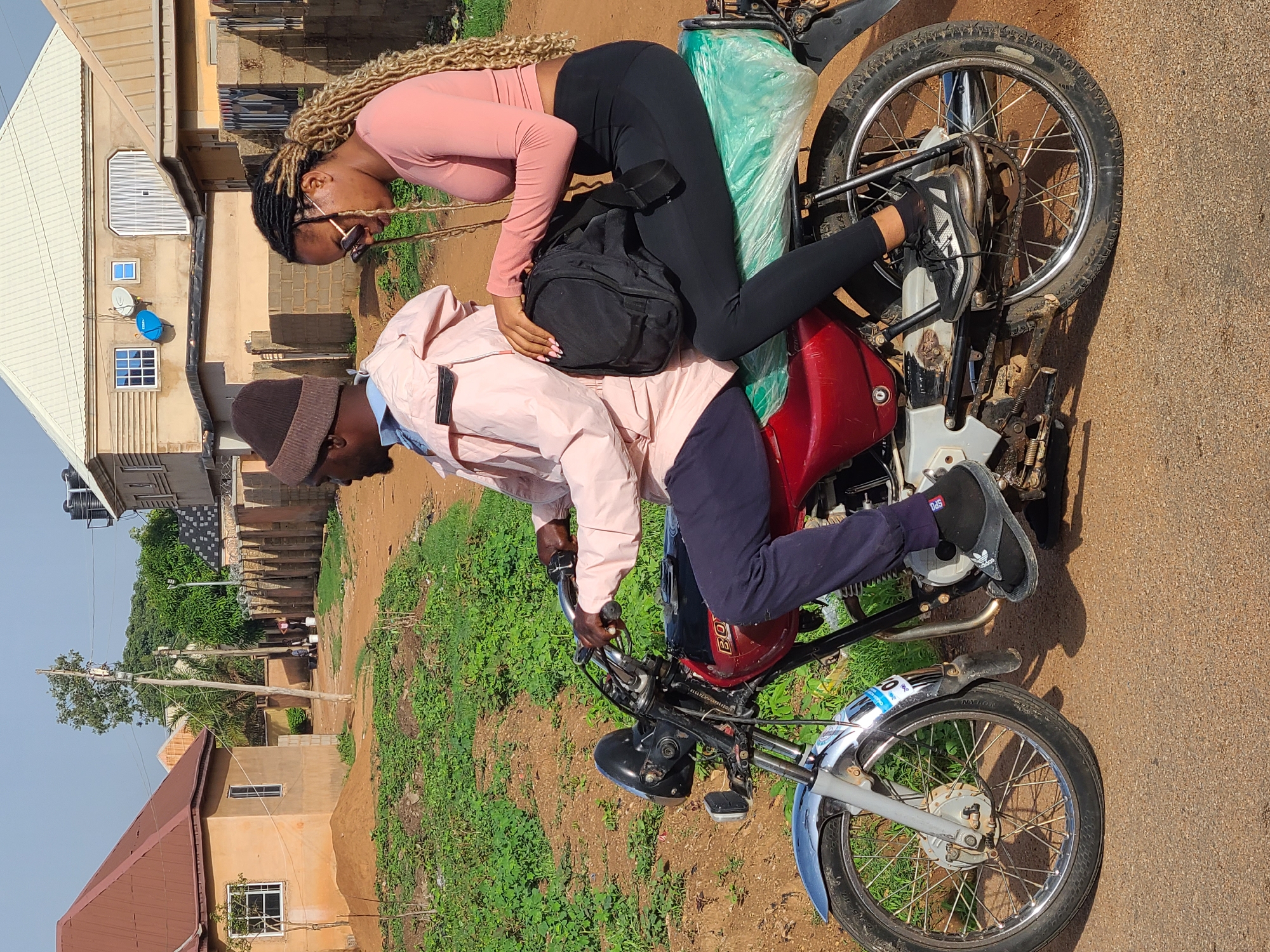 A man and a woman are positioned on a motorcycle on a dirt road in a residential area. The man is seated in the driver's position, wearing a brown hat, a light pink jacket, and blue pants. He's holding the handlebars of the motorcycle. The woman, dressed in a pink long-sleeved top and black leggings, is leaning forward with one arm around the man's shoulders, her body positioned towards the front of the motorcycle. A black backpack is placed between them. The motorcycle is red with silver accents. Behind them, there is a green plastic-covered object strapped to the back of the motorcycle. The surroundings include houses, a dirt path, and green vegetation.