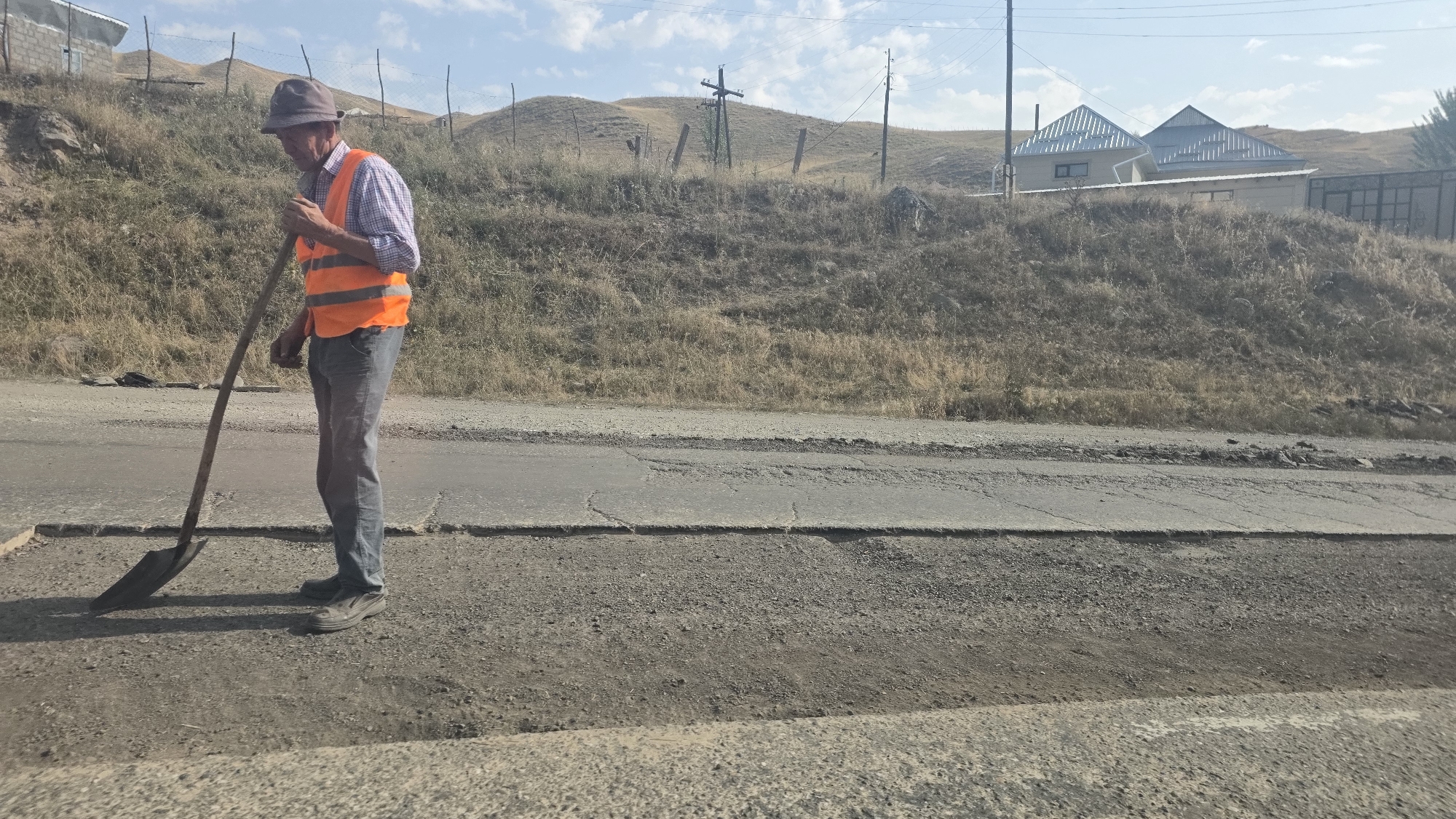 An elderly man wearing a gray hat and an orange safety vest stands on a road, holding a shovel. He is dressed in a collared shirt and jeans. The man appears to be working on the road, which shows signs of disrepair with cracks and uneven surfaces. In the background, there are hills covered with dry grass, a few wooden fence posts, and power lines. Further in the distance, two houses with gray, pyramid-shaped roofs are visible on a slope. The sky is blue with some scattered clouds. The scene suggests an outdoor work setting in a rural or semi-rural area, possibly on the outskirts of Taldyk, Kyrgyzstan.