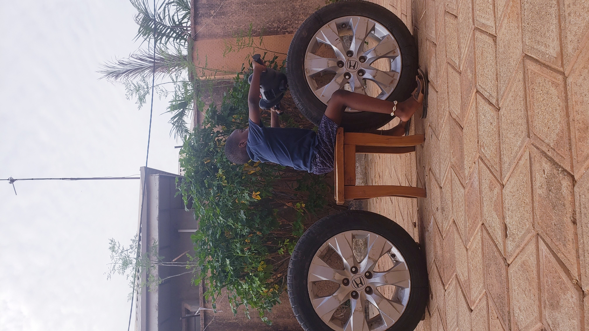 An outdoor, mid-day scene captures a young child engrossed in play. The child, likely a boy, wears a dark blue t-shirt and patterned shorts, and is seated on a simple wooden chair. He holds a black, possibly toy, power tool or object above his head with both hands, suggesting active engagement. A decorative beaded anklet is visible on his right ankle.
Surrounding the child are two car wheels, fitted with silver alloy rims, each prominently displaying the "Honda" logo in its center. One wheel rests vertically behind the child, while the other is positioned in the foreground to his left. The ground is covered with light brown, rectangular paving stones arranged in a herringbone or zigzag pattern. In the background, lush green foliage, including bushes and palm fronds, provides a natural backdrop. Behind the greenery, a light-colored building wall is visible. The upper left portion of the image shows an expanse of bright, slightly overcast sky, with a tall, thin antenna or pole extending upwards from a structure below. The lighting indicates it is daytime. The overall setting appears to be a courtyard or open outdoor area, consistent with Narayi, Nigeria.