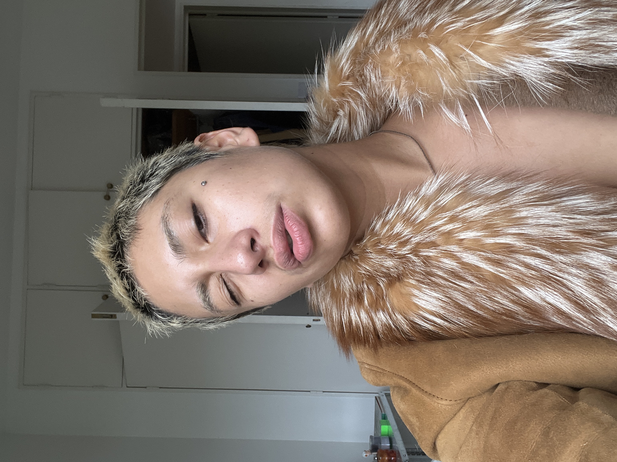 A person with a fair complexion, short, dyed blonde hair, and a stud piercing above their eyebrow is wearing a fur-lined jacket, possibly made of suede or a similar material. The person is tilting their head, with closed eyes and slightly pursed lips. The background shows a plain white door, and some miscellaneous items on a shelf are partially visible in the lower right corner. There is no information in the image to indicate it was taken in Buenos Aires.