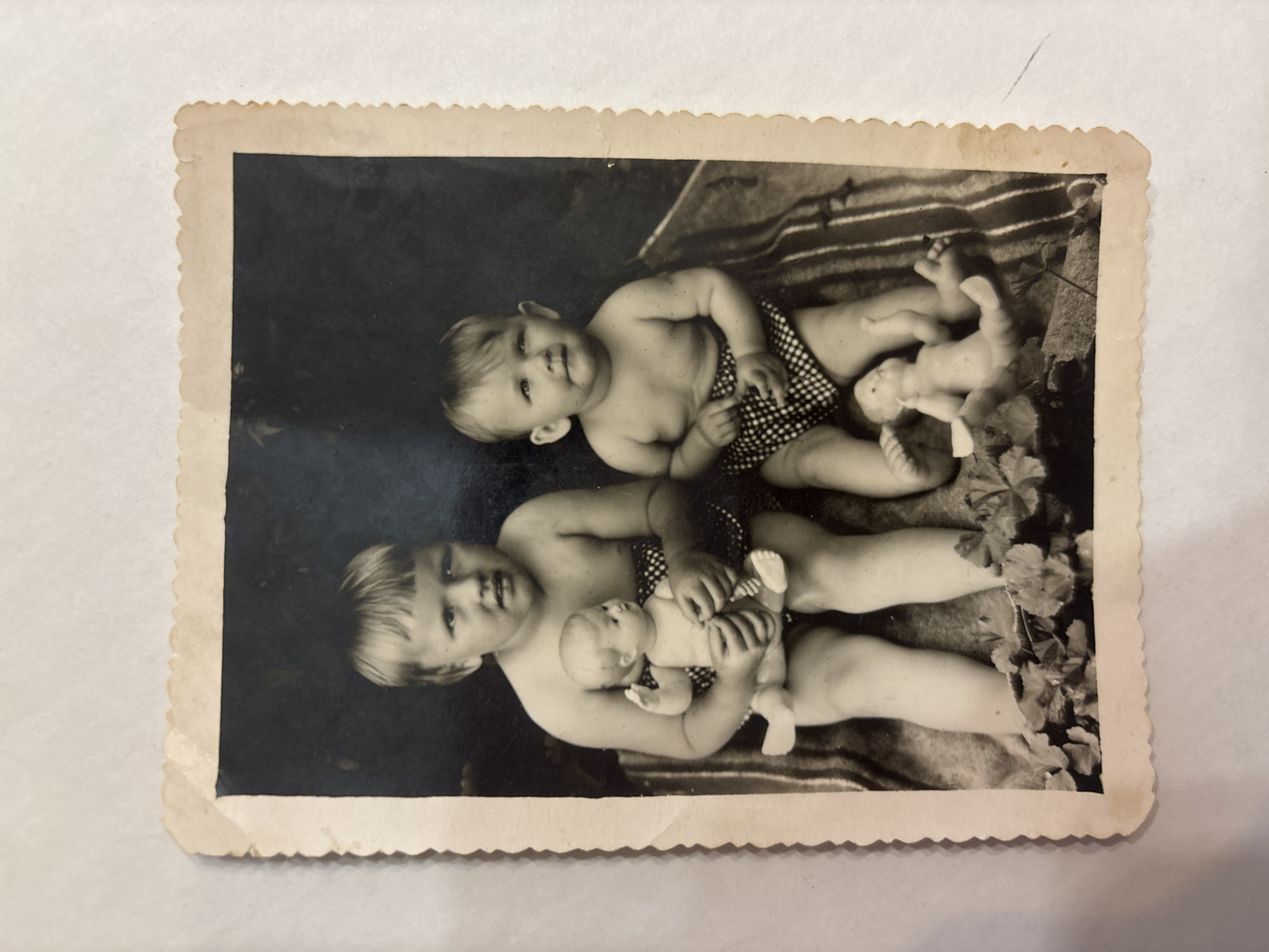 This is a black and white photograph of two young children, likely siblings, posing for a portrait. The older child, appearing to be a toddler, is lying on their back, propped up on one arm, with the other arm holding a doll. They are wearing a patterned romper. Next to and slightly behind them, another baby is also lying down, wearing a similar patterned romper. Both children appear to be in good spirits, with the older child looking directly at the camera with a slight smile. The setting appears to be outdoors, possibly in a garden, with foliage visible in the background and what looks like a striped blanket or rug beneath the children. The photograph has a slightly aged appearance, with a decorative, scalloped border characteristic of older prints. The overall mood is warm and familial, capturing a sweet moment between the two young children. No visible text is present in the image.