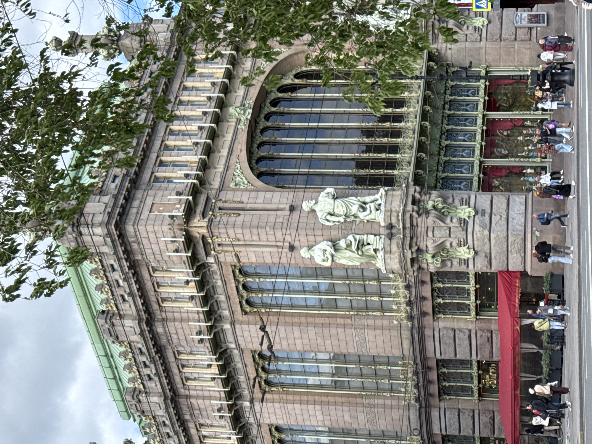 The image shows the exterior of the Eliseyev Emporium building in Saint Petersburg, Russia. The building, characterized by its ornate Art Nouveau architecture, features a stone facade with large arched windows. Sculptures and decorative elements embellish the exterior. A green roofline is visible at the top left of the image. On the ground level, several people are walking along the sidewalk in front of the building, some standing near the entrance, which is covered by a red awning. Foliage from trees obscures the top of the building and the sky in the background. A street sign is also visible near the top.