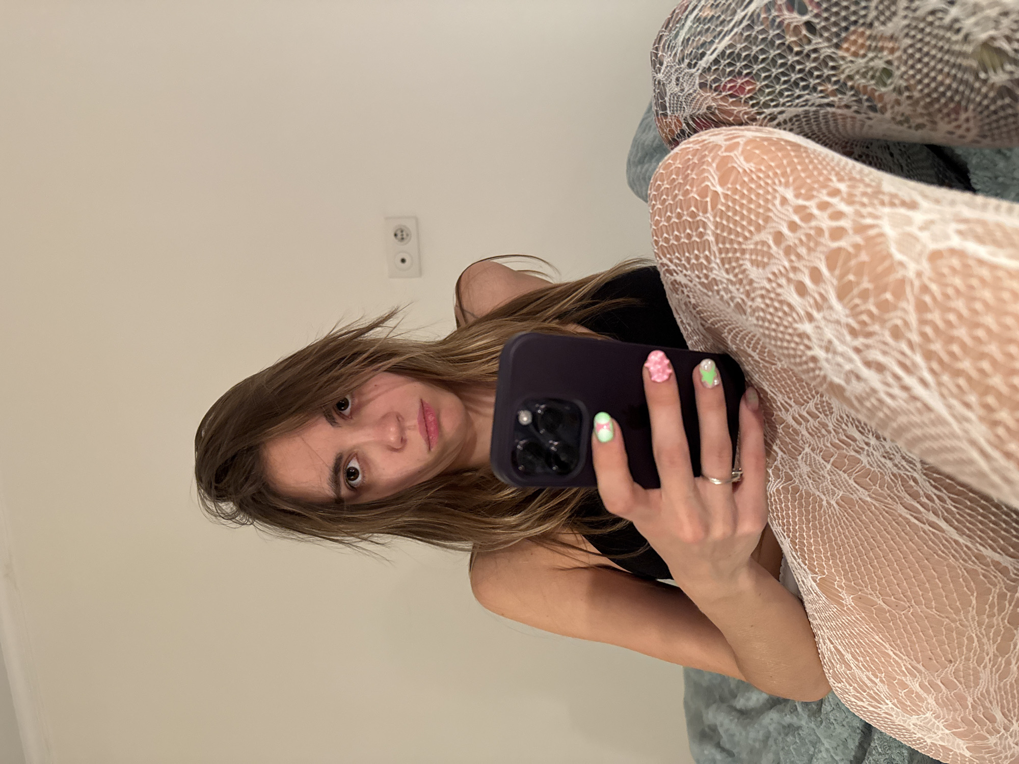 A young woman is taking a selfie in a room with light-colored walls. She is lying down, holding a smartphone in her right hand, which is angled to capture her face and upper body. She has long, brown hair that is slightly messy, framing her face. Her eyes are wide open, and she is looking directly into the camera. Her lips are slightly parted, and she appears to have light makeup on. She is wearing a black top and white lace stockings. Her nails are painted with colorful designs, including pink flowers and green accents. A silver ring is visible on her right ring finger. The room is simply furnished, with a white electrical outlet visible on the wall behind her. The overall lighting suggests it is daytime. The image is a close-up, focusing on the woman and her immediate surroundings.