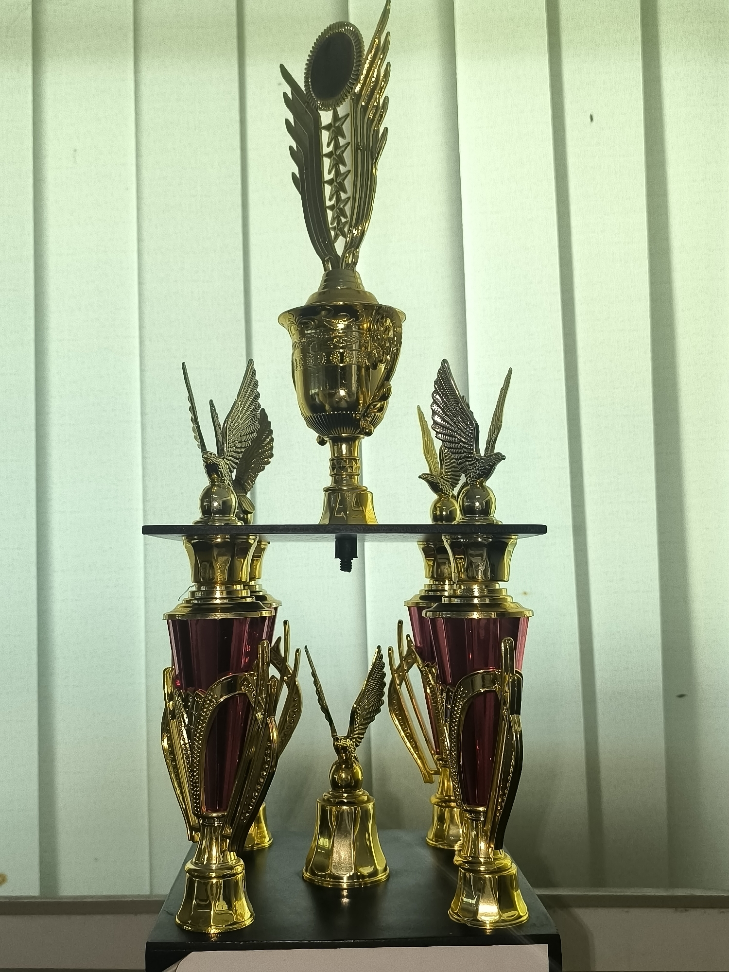 A tiered trophy display is centered in the frame, set against a light green backdrop of vertical blinds. The trophies are primarily gold, with some featuring deep red accents. The arrangement includes a large central trophy at the top, adorned with wings and stars. Below this, on a dark shelf, sit two smaller gold trophies, each topped with a winged figure. The lower level showcases two larger, more elaborate trophies with red cylindrical cups framed by gold accents. In front of these, and slightly lower, is a small, bell-shaped gold trophy. The display rests on a dark surface, which in turn is on a lighter surface at the bottom of the frame. The lighting is soft, casting subtle shadows. The image was taken in Kalabbirang, Indonesia.