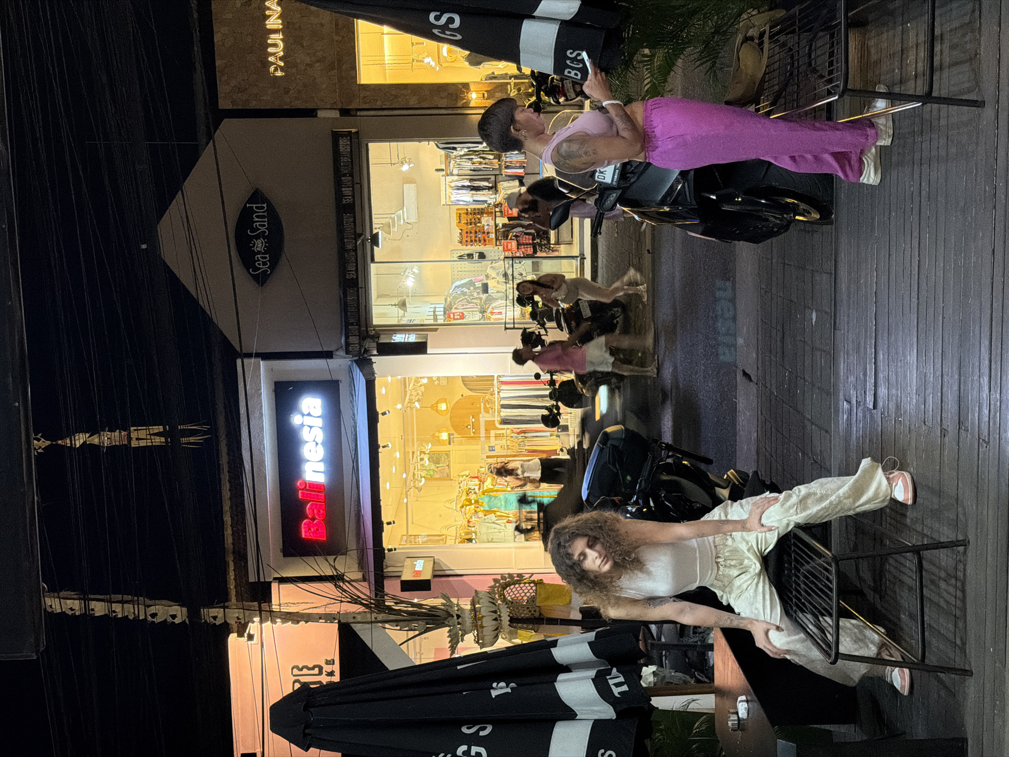 The image captures a lively street scene in North Kuta, Indonesia, at night. In the foreground, a woman with curly hair sits on a metal chair, wearing a white top and light-colored pants. Her pose suggests relaxation, with one hand resting on her leg. Behind her, a man in a pink tank top and vibrant purple pants leans against a motorcycle, looking at his phone. The background reveals the interiors of several shops, illuminated by warm lights. One shop prominently displays clothing racks filled with garments. The signage visible includes "Balinesia," "Sea Sand," and "PAULINA," indicating commercial establishments. Umbrellas are set up outside some of the shops, suggesting an outdoor seating area. The overall atmosphere appears to be that of a bustling street with people enjoying the evening.