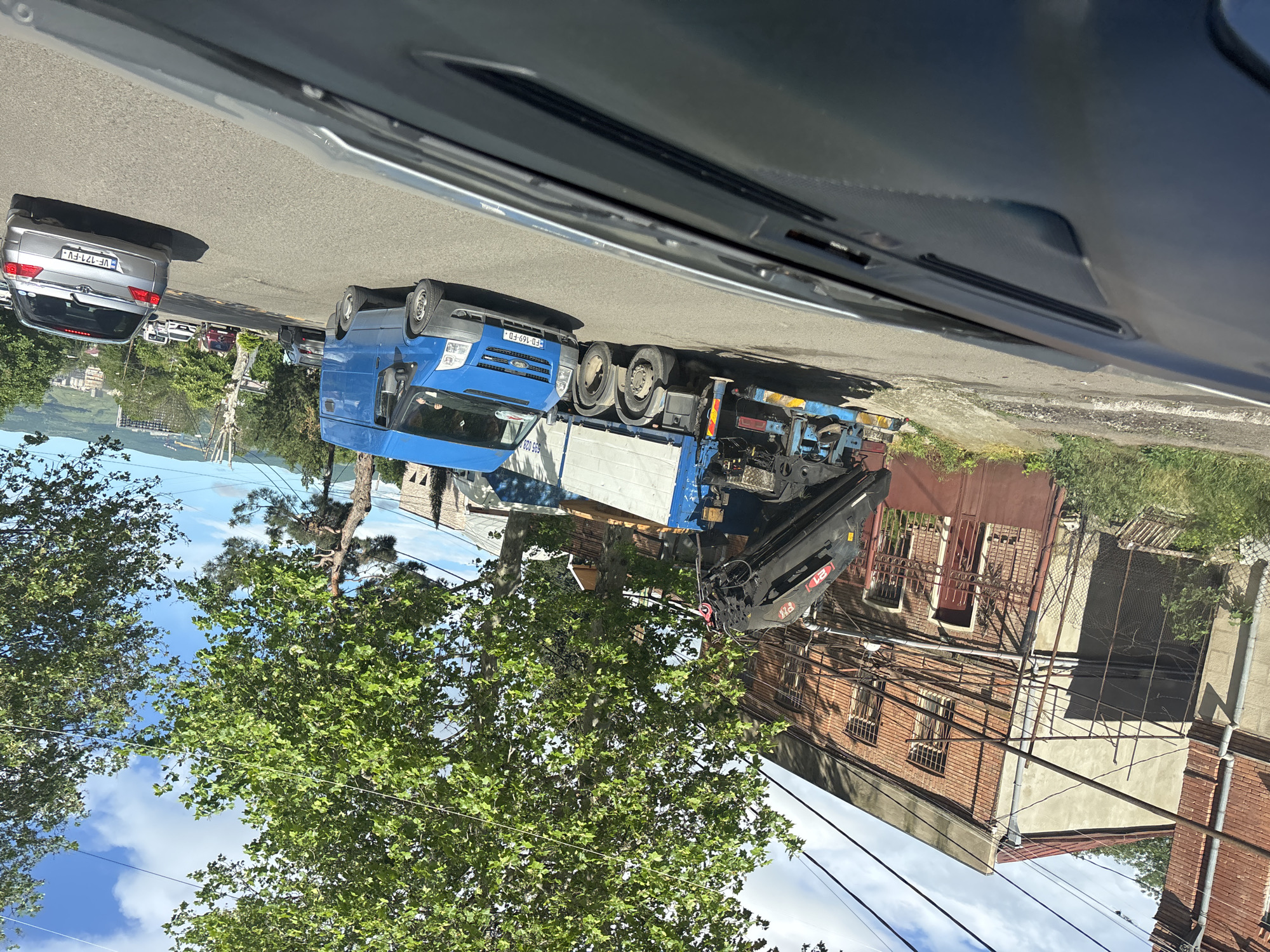 The image shows a blue Ford Transit crane truck parked on a street in Tbilisi, Georgia. The license plate of the truck is FD-169-FD. A gray sedan with license plate VF-121-FV is parked behind the truck. Power lines run along a two-story brick building. Trees and sky are visible. The photo was taken from inside a vehicle.