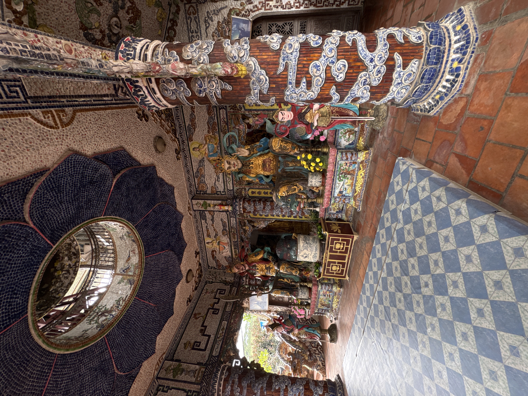 This image depicts the interior of a Buddhist temple, likely Linh Phuoc Pagoda, in the city of Xuân Thành, Vietnam. The temple is elaborately decorated with mosaics made of ceramic shards and glass. In the center of the scene are two large, seated statues, presumably of Buddhist deities or figures of importance. The statue on the right is adorned in gold and green robes, holding a weapon. To its left is another statue, also richly decorated. In front of the statues, there is a small, ornate cabinet.
The architecture is characterized by intricately tiled columns and walls. One prominent column on the right is covered in mosaic patterns featuring blue and white swirling motifs and dragon-like figures. The ceiling features a large, circular opening with a railing, allowing light to filter in from above, and is also covered in mosaic work, resembling lotus petals. The floor is tiled with reddish-brown tiles, with a raised platform covered in a blue and white checkered mat in the foreground. The walls are adorned with colorful murals and smaller mosaic figures. The overall impression is one of opulence and detailed craftsmanship typical of Vietnamese Buddhist temples. The lighting suggests it is daytime, with natural light coming from the opening in the ceiling and possibly from an unseen entrance.