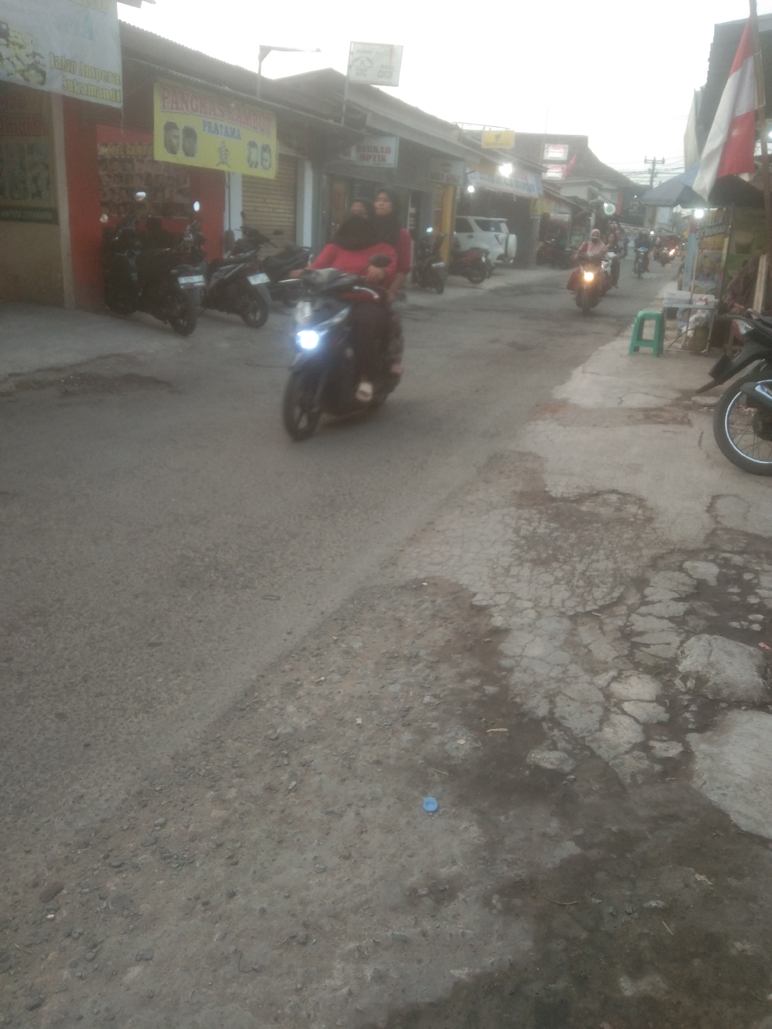 This is a photograph taken on a street in Subang, Indonesia. The scene depicts a residential or commercial area with buildings lining the street. Several motorcycles are visible, with some in motion and others parked. In the foreground, a motorcycle is prominently displayed with its headlight illuminated, suggesting it is either moving or has recently been in use. The road surface appears to be asphalt, with some signs of wear and tear, including cracks and patching.
In the mid-ground, a person is riding a motorcycle, and another individual is seated behind them. The rider is wearing a red top and a dark headscarf, while the passenger also wears dark clothing and a headscarf. They appear to be in motion, moving down the street. Further down the road, other motorcycles and people are visible, indicating a moderately busy street.
The buildings on either side of the street house various establishments. On the left, there appears to be a shop with signage that includes text in Indonesian, possibly indicating a business name or service. On the right, more buildings are visible, with some displaying commercial signage. A green stool is placed on the sidewalk, and a blue plastic bottle lies on the ground near the foreground. The overall lighting suggests it is either early morning or late afternoon. The environment appears to be a typical urban or semi-urban street in Indonesia.
