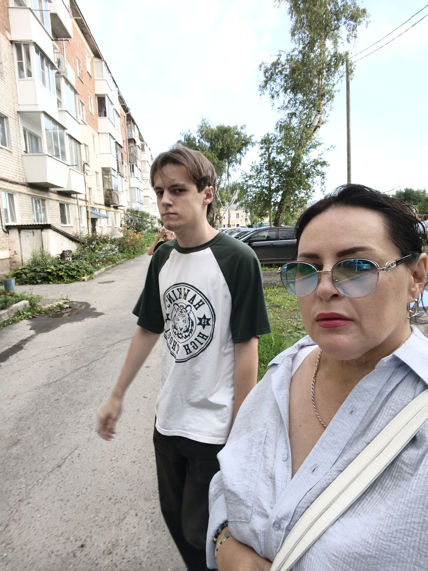 A young man, appearing to be in his late teens or early twenties, walks away from the camera, his head turned back to look over his shoulder. He is wearing a white t-shirt with green sleeves and a circular graphic on the front, and dark pants. To his right and closer to the camera, a middle-aged woman is also walking, facing the camera. She is wearing a light blue collared shirt, round sunglasses with a reflective, blue-tinted lens, and a silver chain necklace. A white strap from a bag is visible across her chest. The background shows a multi-story brick apartment building with white balconies and windows. A paved walkway or road is in the foreground, with some greenery and plants along the side. Several cars are parked in the distance. The scene appears to be in a residential area of Irbit, Russia, given the architecture of the building and the style of the vehicles. The lighting suggests it is daytime, likely afternoon, under a partly cloudy sky.