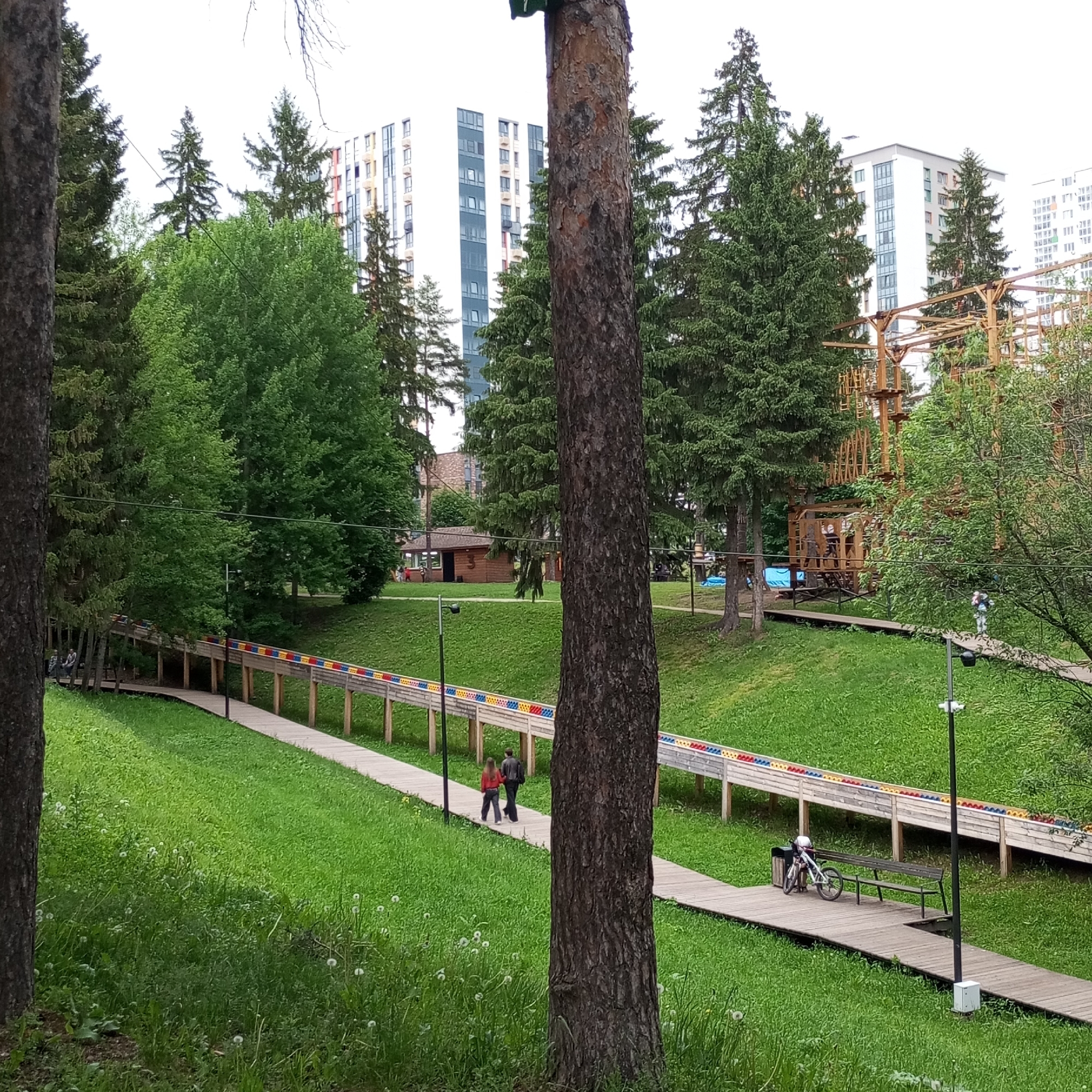 This image captures a park scene in the S.M. Kirov Park of Culture and Leisure in Izhevsk, Russia. The foreground is dominated by the out-of-focus trunks of two large trees, one on the far left and one centrally positioned on the right, framing the view. The ground consists of a sloping expanse of green grass interspersed with scattered white dandelions.
In the midground, a light-colored wooden walkway or ramp, supported by numerous wooden posts, extends from the left and gently curves towards the right, ascending slightly. Along the right side of this walkway, a low wooden barrier features a continuous pattern of small, repeating red, blue, yellow, and green rectangular segments. On the walkway, near the right foreground tree, a white bicycle with a light-colored helmet resting on its handlebars is parked beside a dark park bench and a dark trash receptacle. A tall, dark lamppost with two light fixtures is situated on the walkway between the bench and a nearby smaller tree. Two individuals, a woman in a red top and dark pants walking slightly ahead of a man in dark clothing, are visible walking away from the viewer on the walkway, positioned behind the right foreground tree. Further along the walkway to the left, two to three additional indistinct figures are present.
The background is densely populated with a variety of trees, including both tall conifers and leafy deciduous trees. Through and above the tree line, several modern multi-story residential buildings are visible, characterized by white or light grey facades, numerous dark-paned windows, and some colored architectural accents in red, blue, and yellow. On the far right, partially obscured by trees, a large wooden structure typical of an adventure or rope park, consistent with the "Vysotnyy Gorodok" (High-Altitude Town) attraction, rises with multiple platforms, ladders, and bridges. Near the center-left, a small, dark-roofed wooden building is nestled amidst the trees. The sky is uniformly overcast and white.