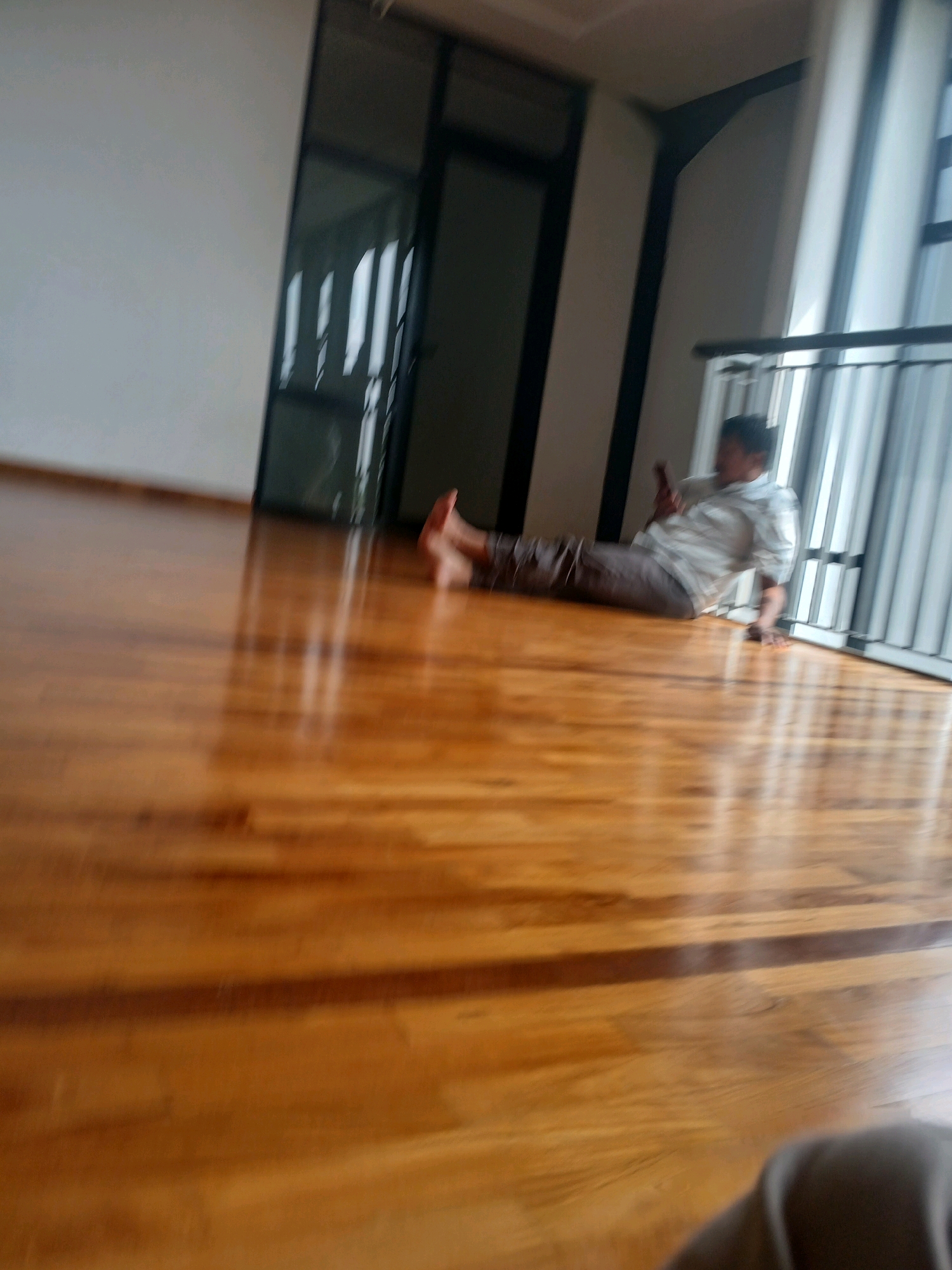 A person is sitting on a polished wooden floor in what appears to be a modern, spacious room. The person is wearing a light-colored, patterned shirt and dark pants, and they are holding a dark object, likely a phone, in their right hand. Their legs are extended in front of them, and they are leaning back on their left arm. The room has large windows with vertical blinds that are letting in natural light, creating reflections on the floor. To the left of the person, there is a dark doorway or partition. In the background, there is a railing, suggesting the area might be an elevated walkway or balcony. The lighting in the room is bright, highlighting the sheen of the wooden floor. The overall scene suggests a moment of relaxation or casual activity. The image lacks specific architectural details that would definitively place it in Arjasari, Indonesia, but the interior design is consistent with contemporary residential or commercial spaces found in urban or suburban areas.
