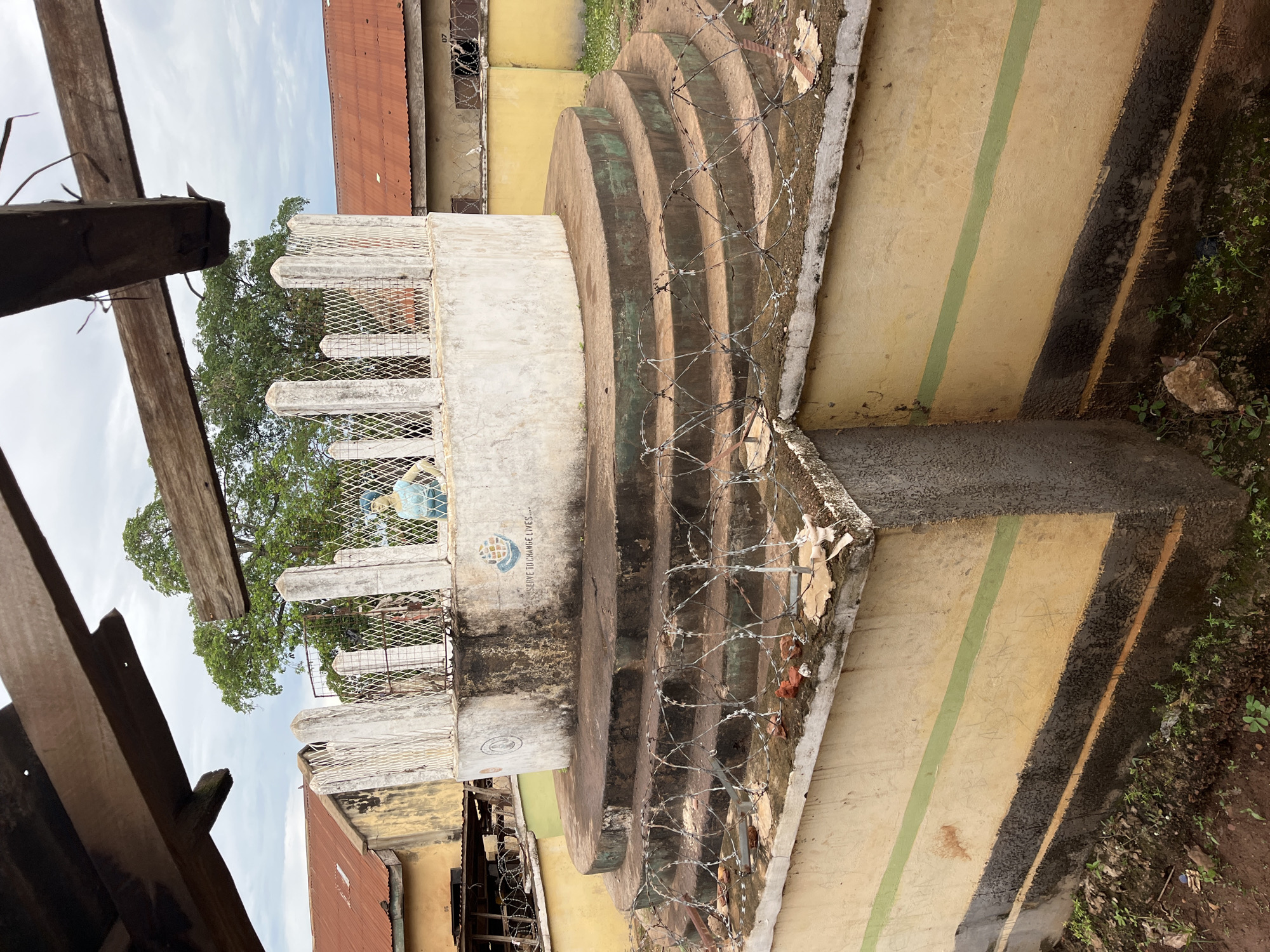 This image depicts an outdoor scene in Idah, Nigeria, centered around a multi-tiered concrete structure. The structure comprises a square or rectangular base, painted light yellow with a horizontal green stripe, and a lower dark-stained section with a thinner orange stripe. Rising from this base are three concentric, circular concrete steps, dark and worn, around which razor wire (barbed tape) is irregularly placed.
Above the steps is a circular concrete platform, topped by a thinner circular wall of light gray/white concrete, exhibiting dirt and staining. This upper wall is enclosed by a white mesh fence supported by multiple white concrete posts. Inside this enclosure, a blue and light-colored globe sculpture, depicting the Earth, is visible on the right. On the exterior of the circular wall, a faded blue and white logo featuring a globe and hands is present, alongside the text "...SERVE TO CHANGE LIVES...". Another faint, circular emblem is visible on the left side of the same wall.
In the background, single-story buildings with corrugated reddish-brown metal roofs and faded yellow/ochre walls are visible on both the left and right. The building on the right has a small window-like opening with security bars or mesh, and additional razor wire is present along its lower wall. The number "07" is marked above this opening. A large, verdant tree stands prominently behind the central structure, its canopy extending above the surrounding buildings. The ground surrounding the base of the structure is earth with sparse green vegetation and debris.
Overhead, partially framing the top of the image, are two rough-hewn wooden beams, showing signs of age and splintering, consistent with a damaged or incomplete wooden framework. The sky is overcast.