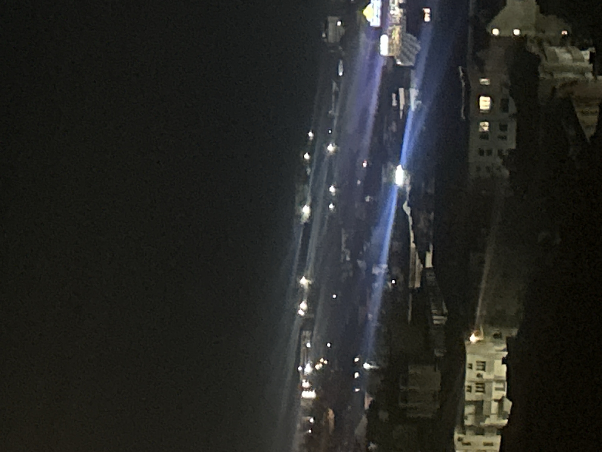 The image displays a night-time elevated view of an urban or suburban landscape in the city of Unknown, Unknown. The upper portion of the frame is a dark, nebulous sky, through which multiple distinct, concentrated beams of light, primarily blue-white, traverse horizontally across the mid-ground. These beams originate from various points below the visible horizon line. Numerous scattered, bright points of artificial light are visible throughout the distant and mid-ground, indicating streetlights, building lights, or vehicle headlights. The foreground is darker, featuring the silhouettes of buildings and trees. Prominent structures include a multi-story, light-colored building on the lower left with several illuminated windows, and another building on the lower right also exhibiting illuminated windows. No individuals, specific actions, or interactions are discernible within the scene. The atmosphere appears hazy or foggy, enhancing the visibility of the light beams.