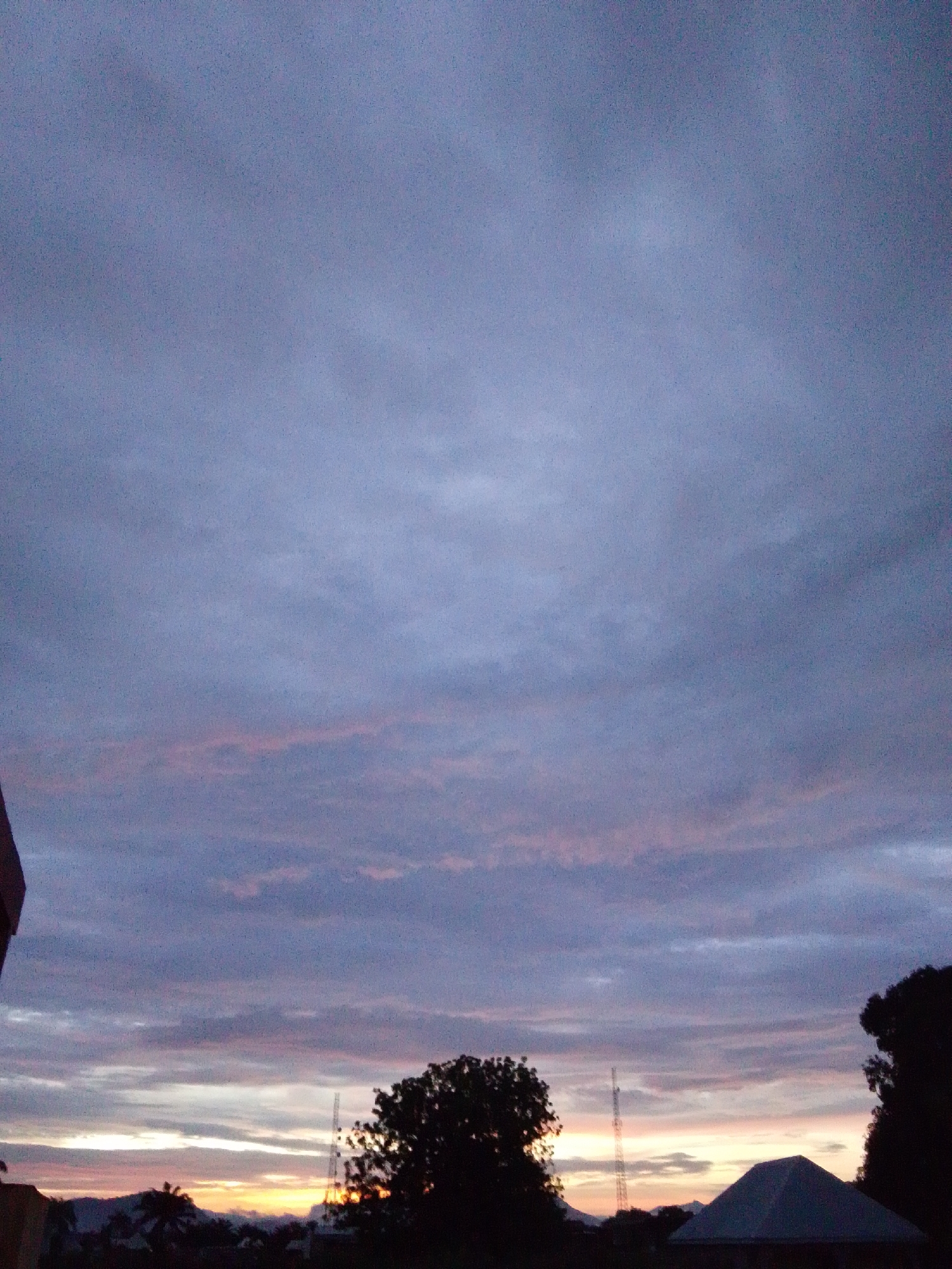 A wide-angle, eye-level, medium shot captures a dusky sky over what appears to be the city of Serti, Nigeria. The sky is predominantly a soft lavender and grey, with streaks of pale pink and orange clouds near the horizon. Silhouetted against this twilight sky are several trees and a conical roofed structure, possibly a building or shelter, in the foreground. A tall, thin tower, likely a communication mast, stands in the mid-ground. The landscape below the horizon is dark and indistinct, with hints of hills and smaller structures. The image is grainy, suggesting it was taken in low light conditions, possibly at dusk or dawn. There are no people or animals visible in the scene.