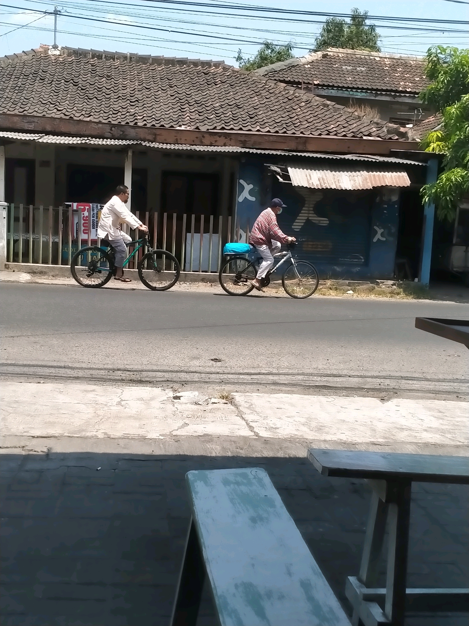Two individuals are riding bicycles on a paved road in front of a row of houses in Kasihan, Indonesia. The house on the left has a tiled roof and a white facade with a wooden fence in front of it. A red and white sign is visible behind the fence. The house on the right is painted blue with white graphics and also has a tiled roof. The road is asphalt and there is a concrete sidewalk or pathway along the side of the road. In the foreground, out-of-focus benches and tables suggest a seating area, possibly an outdoor cafe or dining space. The sky is clear and blue, and there are trees visible in the background. The person on the left is wearing a light-colored long-sleeved shirt and dark pants, while the person on the right is wearing a plaid long-sleeved shirt, white pants, and a dark cap. Both individuals appear to be wearing face masks.
