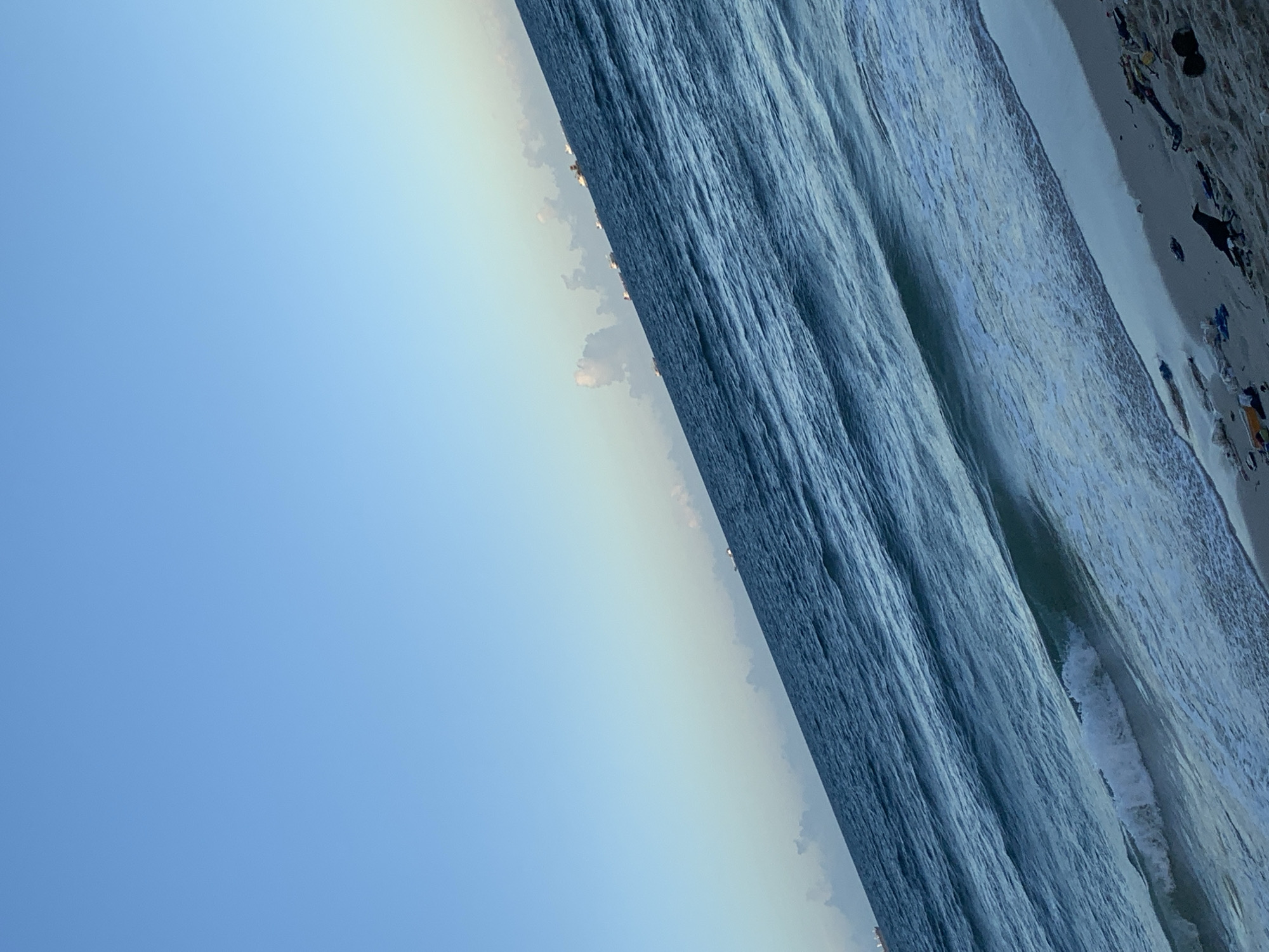 The image displays a coastal landscape, likely captured in Dar es-Salaam, Tanzania, featuring a serene interaction of sky, ocean, and a narrow strip of beach. The scene is primarily divided into a clear blue sky in the upper portion and a vast ocean, with a small section of sandy shoreline visible at the lower right.
The sky occupies the upper left quadrant, presenting a gradient of light blue, brighter and clearer at the top and becoming slightly hazy or lighter towards the horizon line. This horizon cuts diagonally across the image from the top right to the mid-left, where a few wispy, lightly illuminated clouds are visible, suggesting either early morning or late afternoon light.
The majority of the frame is dominated by the ocean, characterized by its deep, dark blue color. The water surface exhibits numerous ripples and visible wave patterns, indicating its dynamic movement. Closer to the shore, particularly in the mid-right, patches of white foam from breaking waves can be seen, emphasizing the active nature of the sea.
At the very bottom right, a narrow band of light-colored sand forms the beach. This sandy area appears somewhat rough, with indistinct dark shapes scattered across it, possibly debris or natural elements washed ashore. No identifiable people, buildings, or specific objects are clearly discernible within the image.
The overall impression is one of natural tranquility and spaciousness. The soft lighting suggests a peaceful time of day. There is no visible text present in the media file.