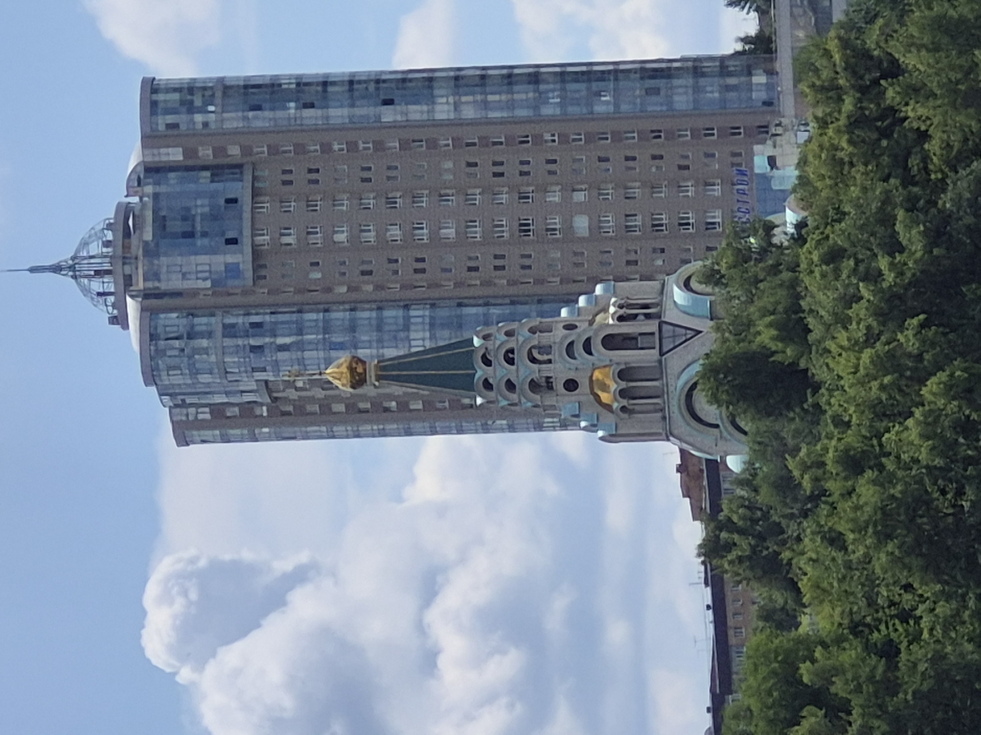 In Samara, Russia, a unique cityscape unfolds where a modern, glass-and-concrete skyscraper stands in stark contrast to an ornate orthodox church with a golden dome. The skyscraper, characterized by its numerous windows and sleek design, is partially obscured by the traditional architecture of the church, which features intricate detailing and vibrant colors. Lush green trees form a dense canopy at the base of the scene, creating a natural foreground that softens the urban environment. The sky above is a mix of blue with large, fluffy white clouds, adding a sense of depth and atmosphere to the image. The juxtaposition of old and new architectural styles, combined with the natural elements, paints a vivid picture of Samara's urban landscape.