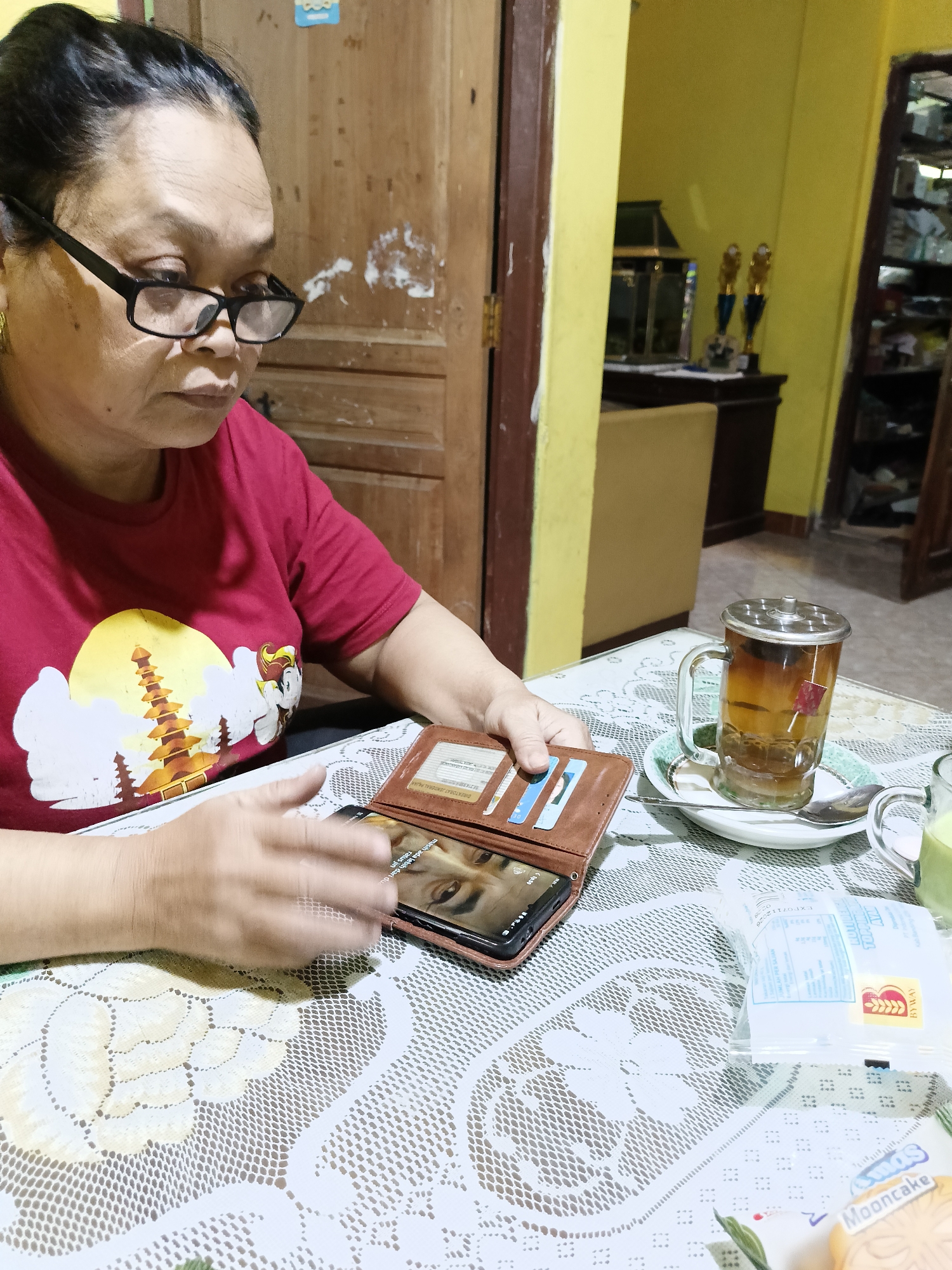 An older woman with dark hair and glasses is sitting at a table in what appears to be a home in Sukorini, Indonesia. She is wearing a red t-shirt with a graphic of a temple on it. In front of her, on a white lace tablecloth, are a brown wallet, a smartphone with a photo of a person on the screen, a glass mug filled with tea, and a plate with a spoon. In the background, a doorway leads to a room with yellow walls. There are shelves with various items, including trophies, in the background. A light yellow chair is partially visible.