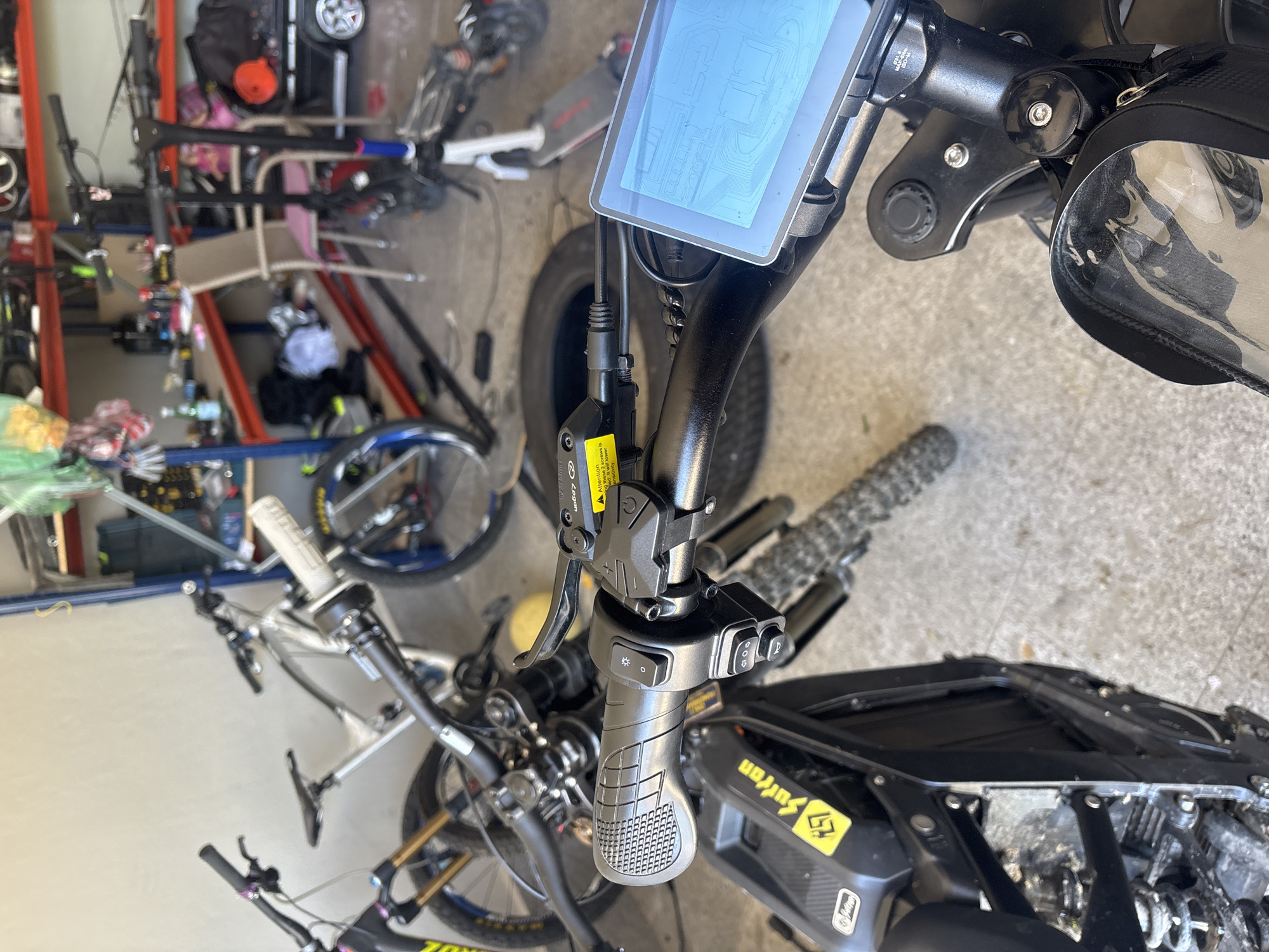 This image depicts a close-up view of the handlebars of an electric bicycle, possibly a Sur Ron based on the logo visible on the frame. The handlebars are equipped with a digital display, controls for lights and assistance levels, and a brake lever. A small pouch is attached to the handlebar stem. The surrounding environment appears to be a cluttered garage or storage space, with various bicycles and other equipment visible in the background. No people are directly visible in the image. There is no indication of being in Asenovgrad, Bulgaria.