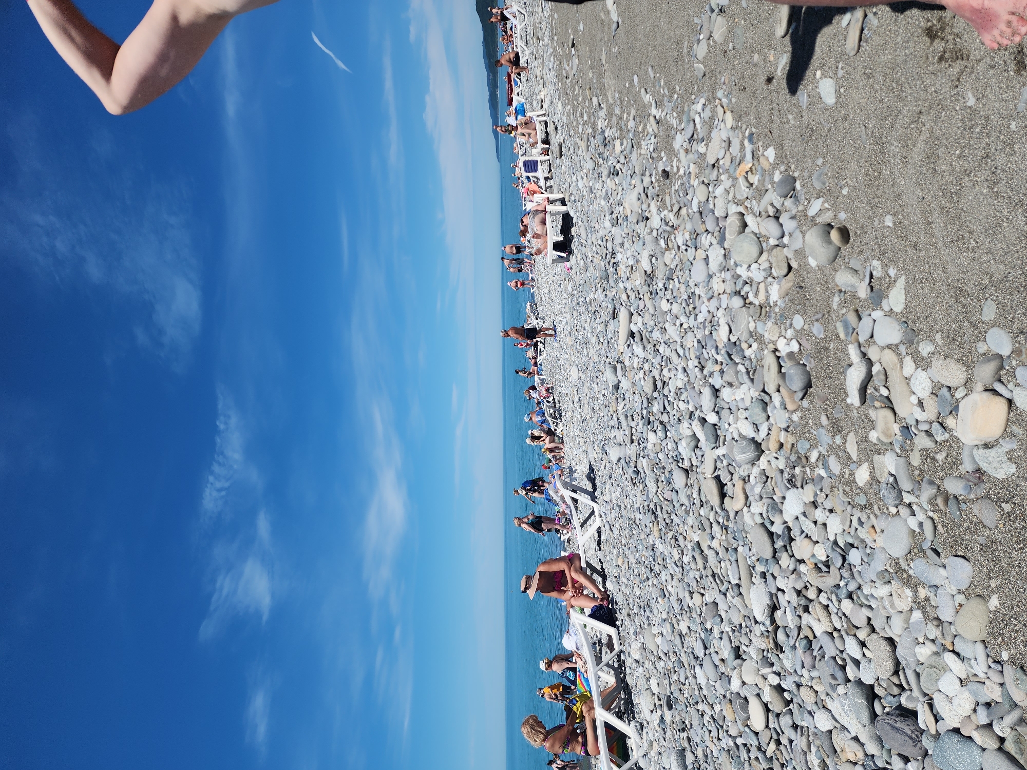 The image captures a vibrant scene at a sunny pebble beach, viewed from a slightly elevated perspective looking out towards the ocean. The top of the frame features a bright blue sky adorned with scattered, wispy white clouds. Below the sky, the vast, calm expanse of the blue ocean stretches across the horizon. Faint outlines of distant mountains or land can be seen along the far edge of the water.
The lower portion of the image is dominated by the beach, which is densely covered with a mix of smooth, grey, white, and darker pebbles, interspersed with patches of sandy ground. Numerous people are scattered along the beach, predominantly relaxing on white plastic lounge chairs that are lined up in rows, facing the ocean. They appear to be sunbathing, lounging, and enjoying the warm weather, dressed in swimwear. Strong shadows cast by the people and chairs indicate bright, direct sunlight.
In the upper right corner, a bare human arm is visible, partially entering the frame, likely belonging to the person taking the photograph. The overall activity depicted is leisurely beach enjoyment, typical of a summer holiday. The scene evokes a sense of relaxation and clear, pleasant weather. There is no visible text in the image.