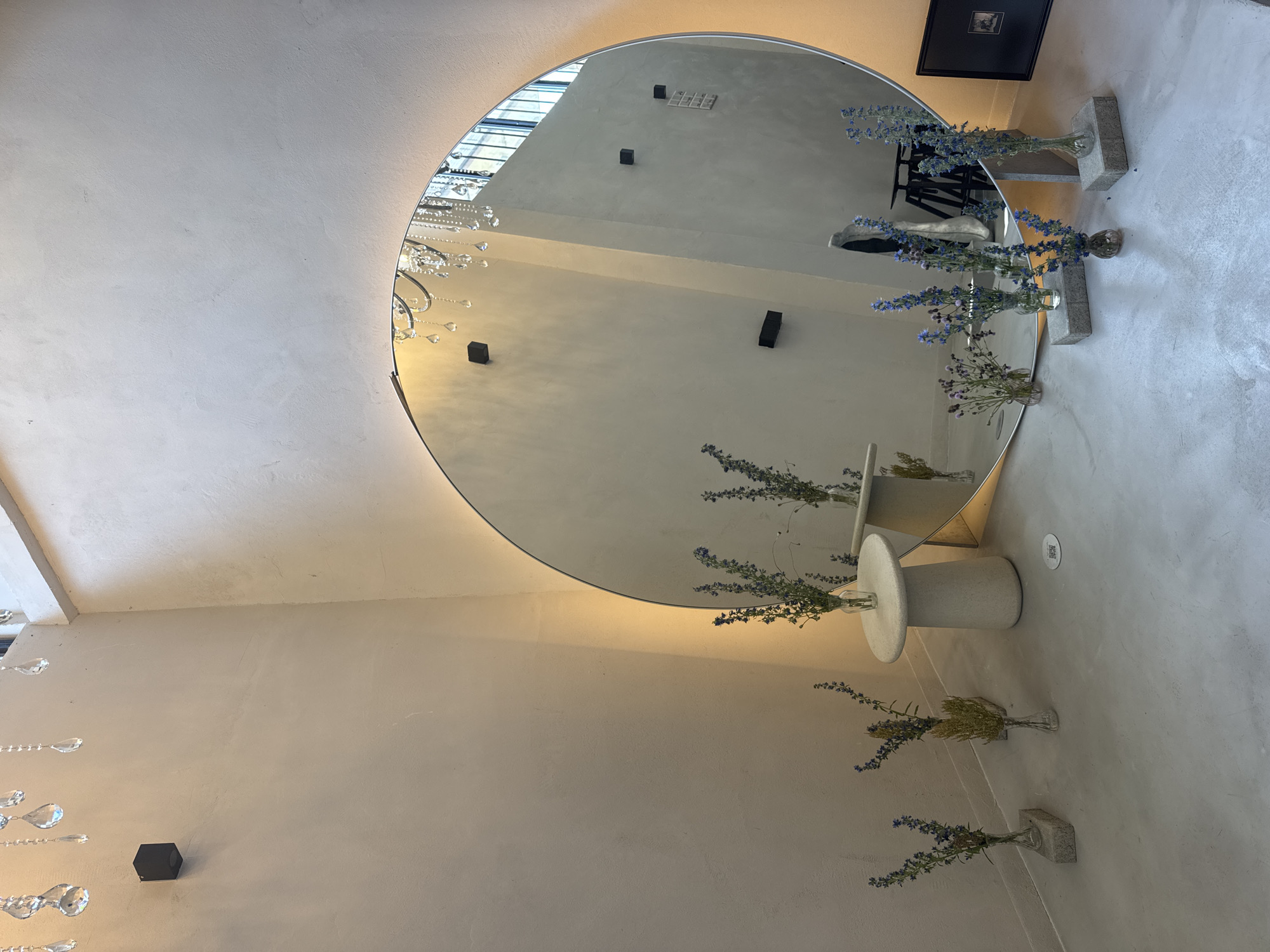 The image depicts an interior space, likely a cafe or a shop, with a minimalist and elegant aesthetic. A large, round mirror dominates the right side of the frame, reflecting the opposite wall and a chandelier with crystal drops. Several small bouquets of dried flowers in glass vases are strategically placed on the floor and a low, round pedestal table. The walls are a light, neutral color with a textured finish. The floor appears to be polished concrete. The overall ambiance is serene and sophisticated, with soft lighting. No people are visible in the scene. The location is identified as Sofia, Bulgaria.