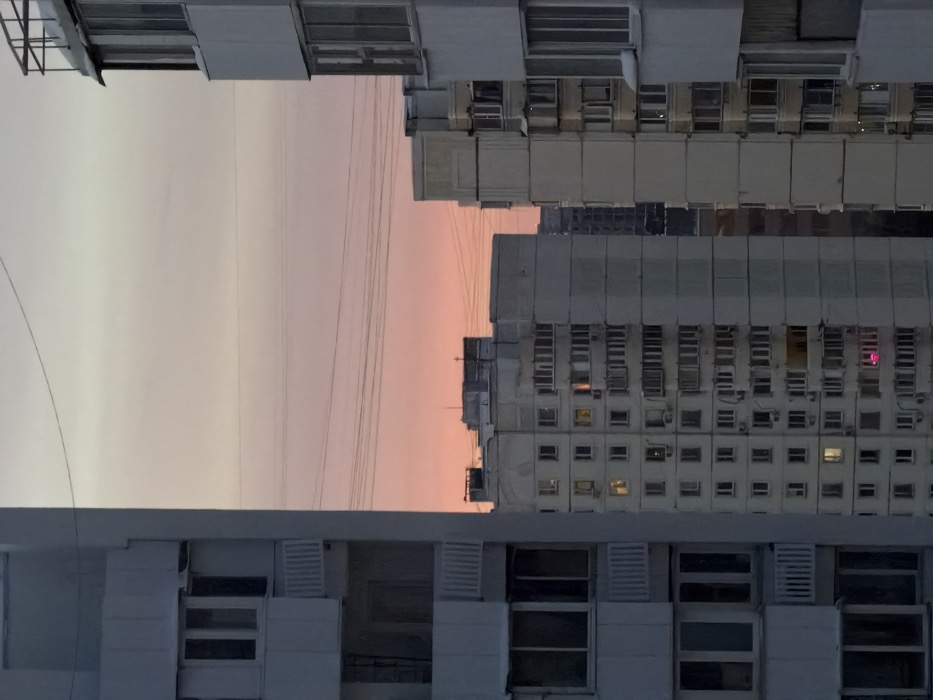 This image displays a residential cityscape in Khimki, Russia, at either dawn or dusk. The scene is framed by two multi-story, light-grey concrete panel residential buildings, one on the left and one on the right, each featuring multiple windows and enclosed balconies. Between these two structures, several additional multi-story residential buildings are visible in the midground. One prominent midground building shows several illuminated windows; most display a warm yellow light, while a singular lower-level window emits a distinct magenta light. Multiple thin, dark horizontal wires traverse the sky across the upper mid-section of the image. The sky transitions from a soft pale orange and pink near the horizon to a lighter, desaturated grey-pink in the upper portion. No individuals are visible within the frame. The architectural style is functional, characteristic of residential blocks.