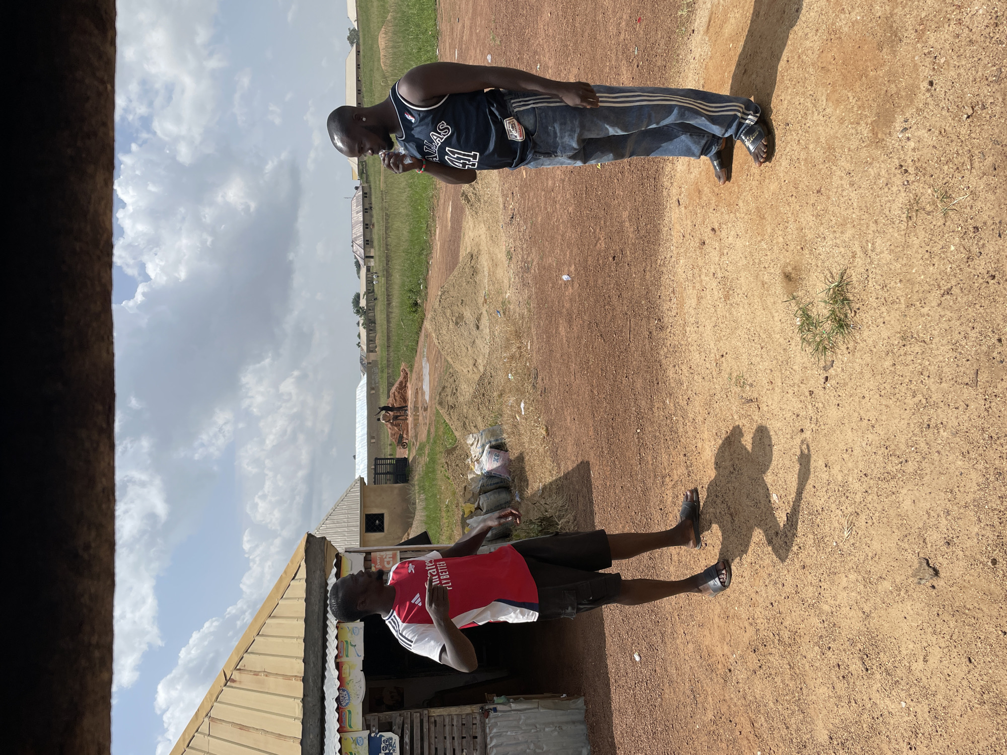 In Wumat, Nigeria, on a sunny day with scattered clouds, two men are standing on a dirt ground. The man on the left, wearing a red and white Arsenal soccer jersey and black cargo shorts, gestures with his right hand. He is standing near a small wooden structure with a corrugated metal roof. To his right, a man in a dark blue basketball jersey with the number "41" and blue jeans stands with his left hand holding a clear plastic bottle to his mouth. Behind them, there are piles of sand, construction materials, and several buildings with corrugated metal roofs. Some people are visible in the distance near the buildings, possibly involved in construction. The environment appears to be an outdoor market or construction site.