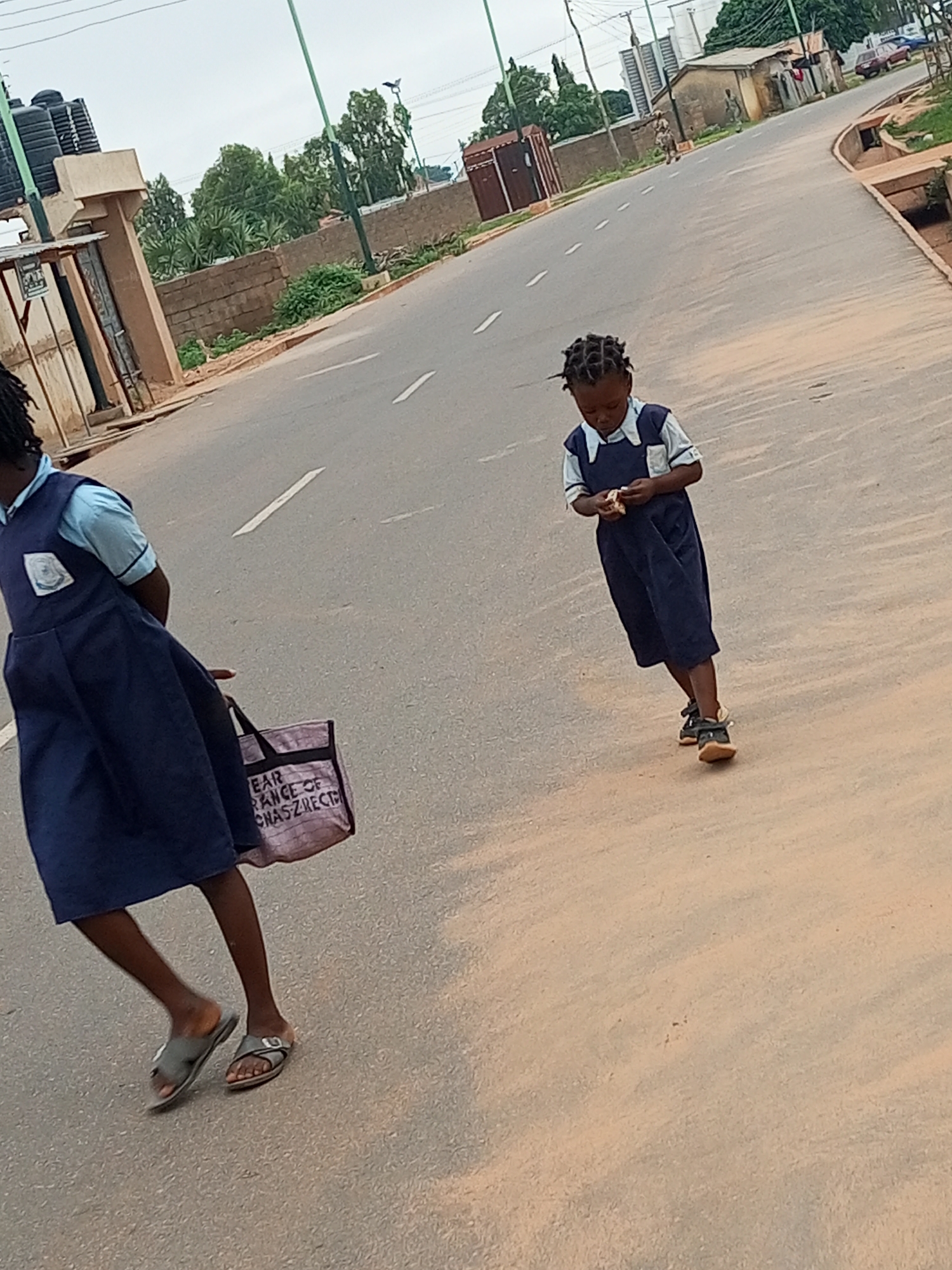 Two young girls, dressed in dark blue school uniforms with light blue collars and sleeves, are walking on a paved road in Tumfure, Nigeria. The girl in the foreground, facing away from the camera, carries a light-colored bag with dark lettering. Her legs are bare below her knees, and she wears grey open-toed sandals. The second girl, walking further down the road and towards the camera, is looking down at something in her hands. She also wears a dark blue school uniform with a light blue collar and sleeves and dark sandals. The road is bordered by a raised concrete curb on the right and a sandy area with some vegetation on the left. In the background, there are brick walls, trees, and buildings, including what appears to be a small structure with a corrugated metal roof. The sky is overcast.