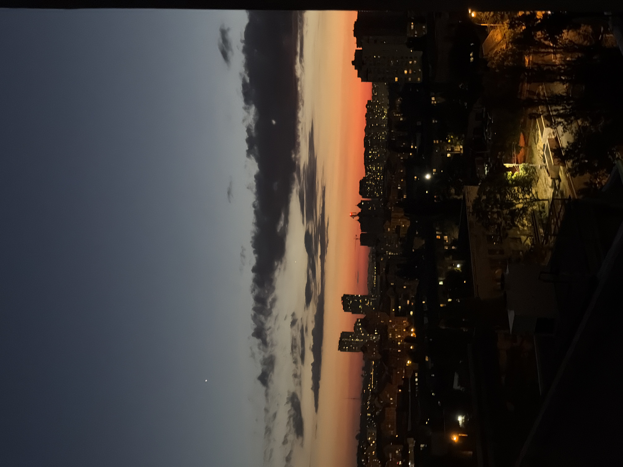 The image is a nighttime aerial view of a city, likely Kyiv, Ukraine, under a dusky sky. The sky transitions from a deep indigo at the top to a vibrant orange and red glow near the horizon, indicating a sunset or twilight. Clouds are scattered across the sky, some dark and heavy, others wispy.
Below the sky, the cityscape is visible, characterized by a dense arrangement of buildings. Many of these buildings have illuminated windows, suggesting that people are inside their homes or businesses. Prominent among the buildings are several high-rise apartment blocks, some with numerous lit windows. The lower parts of the city are darker, with streetlights casting pools of yellow-orange light onto roads and pathways. Trees are scattered throughout the urban landscape, their forms silhouetted against the faint light. The foreground shows a dark, possibly a rooftop, with a thin line of another structure in the distance. There is no visible evidence of people or specific actions occurring in the scene. The overall impression is one of a typical urban environment at dusk, with the city coming to life with artificial light as natural light fades.