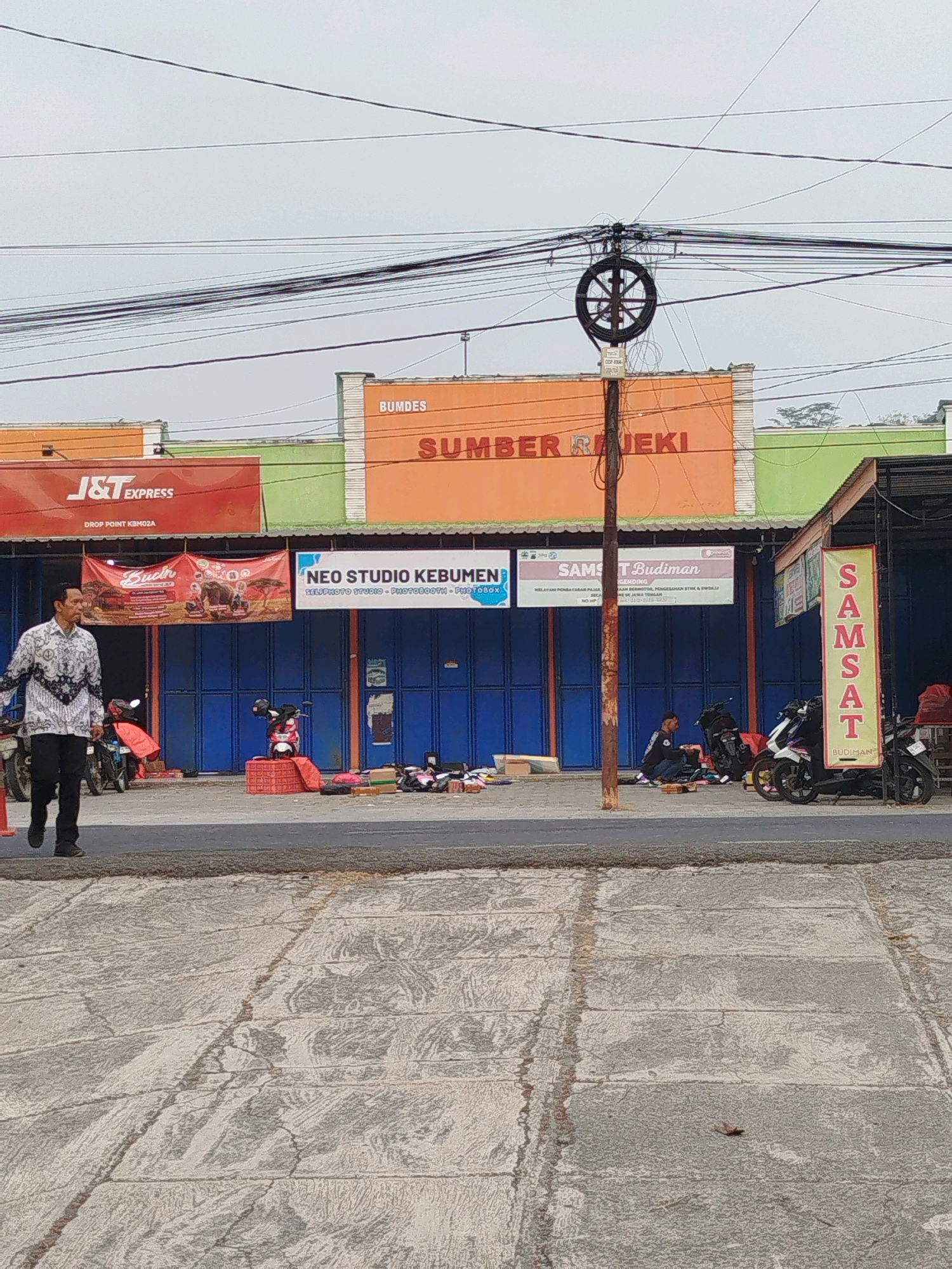 The image shows a street scene in Kedungwaru, Indonesia, in front of a row of businesses. In the foreground, a man in a patterned shirt and dark pants walks from left to right. Behind him, several motorcycles are parked. The buildings in the background have colorful facades and signage. One sign reads "J&T EXPRESS," another says "BUMDES SUMBER REJEKI," and a third advertises "NEO STUDIO KEBUMEN." Further down the row, a vertical banner reads "SAMSAT BUDIMAN." The street itself is paved with asphalt and concrete, with some visible cracks and a raised asphalt section. Overhead, a complex network of electrical wires is visible against an overcast sky. A metal pole with a circular wire spool is also present. Several people are visible sitting or standing near the businesses.