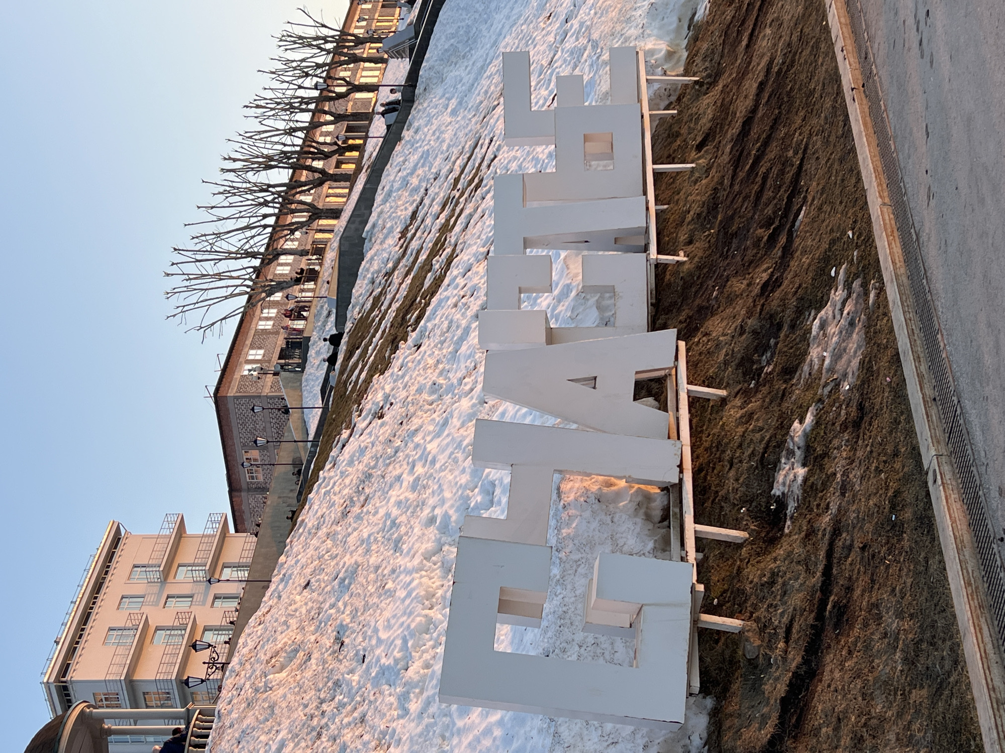 The image depicts a hillside in Perm, Russia, covered with snow and patches of dead grass. The word " счастье " (happiness) is spelled out in large, white, block letters on the hillside. The letters are supported by wooden frames. In the background, buildings are visible, including apartment buildings and a brick structure. The sky is clear and light blue. The sun casts a warm glow on the snow and buildings. The scene appears to be photographed from a low angle, looking up the hillside.