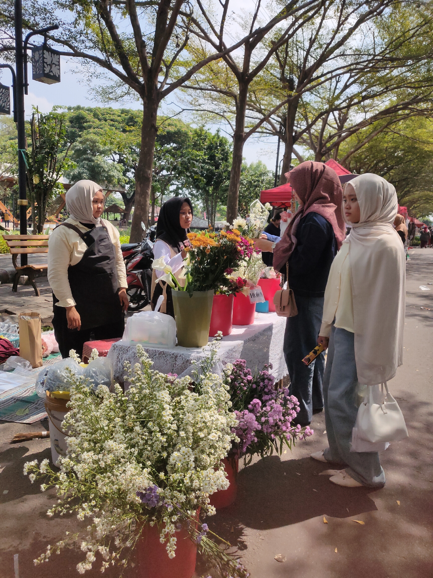 In Ngasem, Indonesia, four women are gathered around a flower stall. The woman on the far left wears a cream-colored hijab, a cream-colored long-sleeved shirt, and a black apron. Next to her, a woman in a black hijab and a white shirt also appears to be working at the stall. Two more women are standing to the right, both wearing hijabs and jeans. The woman on the far right has a cream-colored hijab and a cream-colored outfit, and carries a white handbag. The stall itself is laden with a variety of flowers, including white baby's breath, purple aster, and an assortment of colorful mixed bouquets. Some flowers are arranged in vases, while others are bundled together. Red buckets and a white bucket containing flowers are visible. A price tag of "10.000" is affixed to one of the red buckets. The scene takes place outdoors, under the dappled shade of large trees, with a paved walkway and a park bench visible in the background. The overall impression is of a vibrant outdoor market on a sunny day.