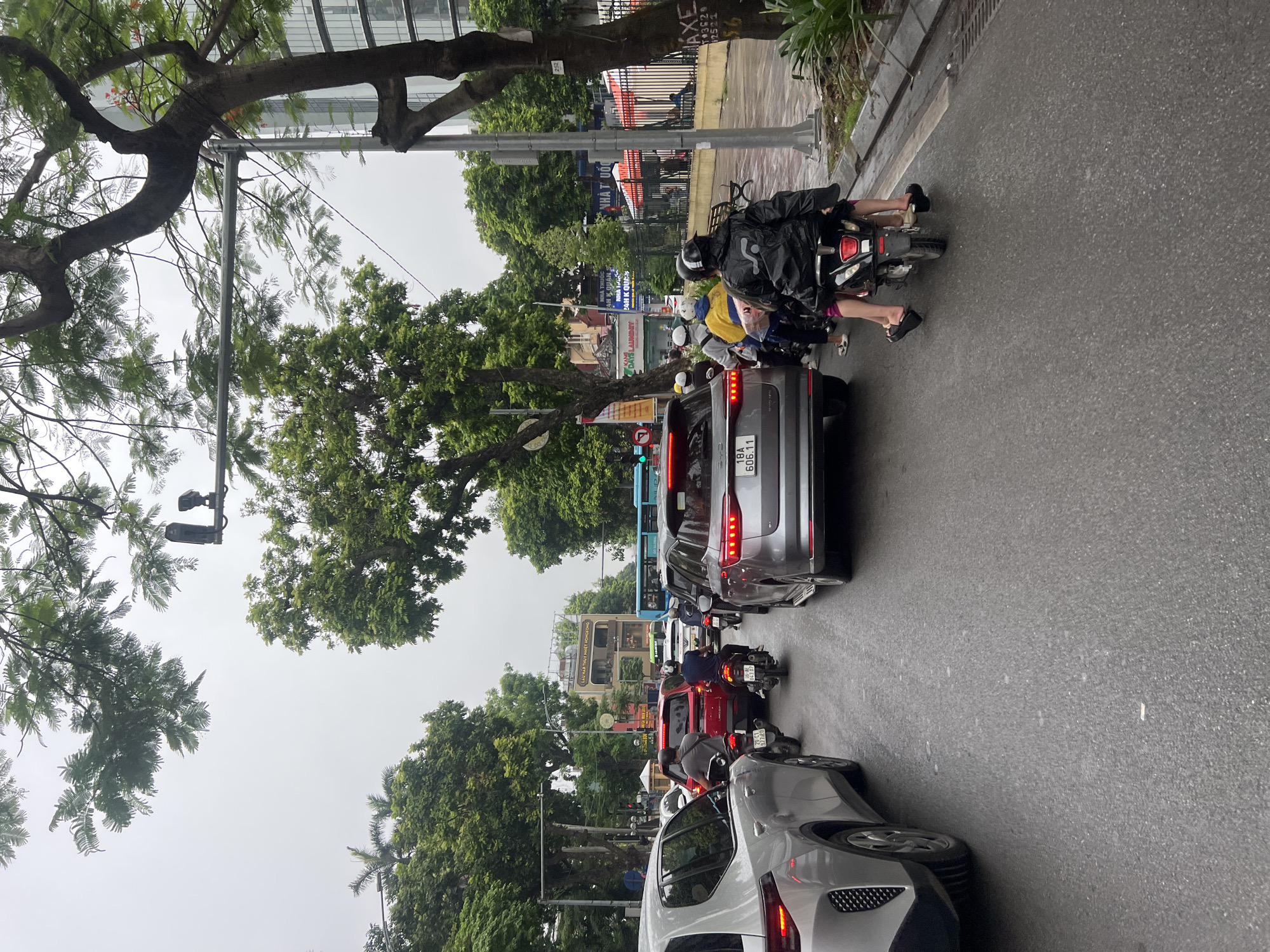 This is a street-level, eye-level, medium shot of traffic congestion on a wet street in Hanoi, Vietnam. The scene is dominated by a line of vehicles, including cars and motorcycles, with trees and buildings lining the sides of the road. The sky is overcast, suggesting a cloudy or rainy day.
In the foreground, on the left, a silver SUV is partially visible. To its right, a silver hatchback is also prominent. In the center of the image, a silver SUV is in the middle lane, with a red car behind it. Several motorcycles are interspersed throughout the traffic. One motorcycle in the right lane has two people on it; the person in the back is wearing a black jacket and purple shorts, and both are wearing flip-flops.
The road appears wet and reflective, indicating recent rain or ongoing precipitation. Trees with lush green foliage line both sides of the street, with some branches extending over the road. In the background, buildings are visible, including a multi-story building with a sign that appears to be in Vietnamese. Traffic signals and streetlights are also present. The overall impression is a typical urban scene in Hanoi, characterized by heavy traffic, abundant greenery, and a bustling atmosphere.