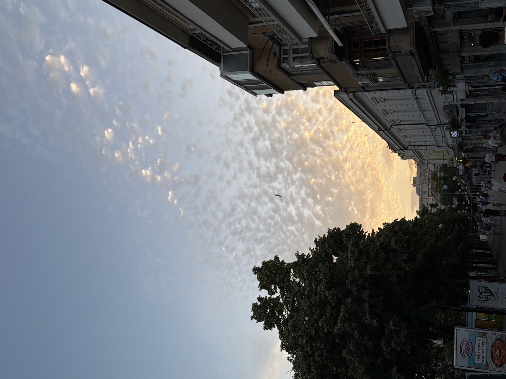 The image shows a street scene in Burgas, Bulgaria, during what appears to be sunset. The sky is filled with a patchwork of light, wispy clouds tinged with orange and yellow hues, indicating the sun is low on the horizon. A large, dark green tree dominates the left side of the frame. On the right, buildings line the street, with architectural details typical of European cities. One building in particular has several balconies and windows, some with ornate frames. The street itself is paved and appears to be a pedestrian area, with a few people walking and gathered. There are posters and signs visible along the street, one of which has text in Bulgarian. The overall impression is a lively urban environment at the end of the day, bathed in the warm glow of twilight.