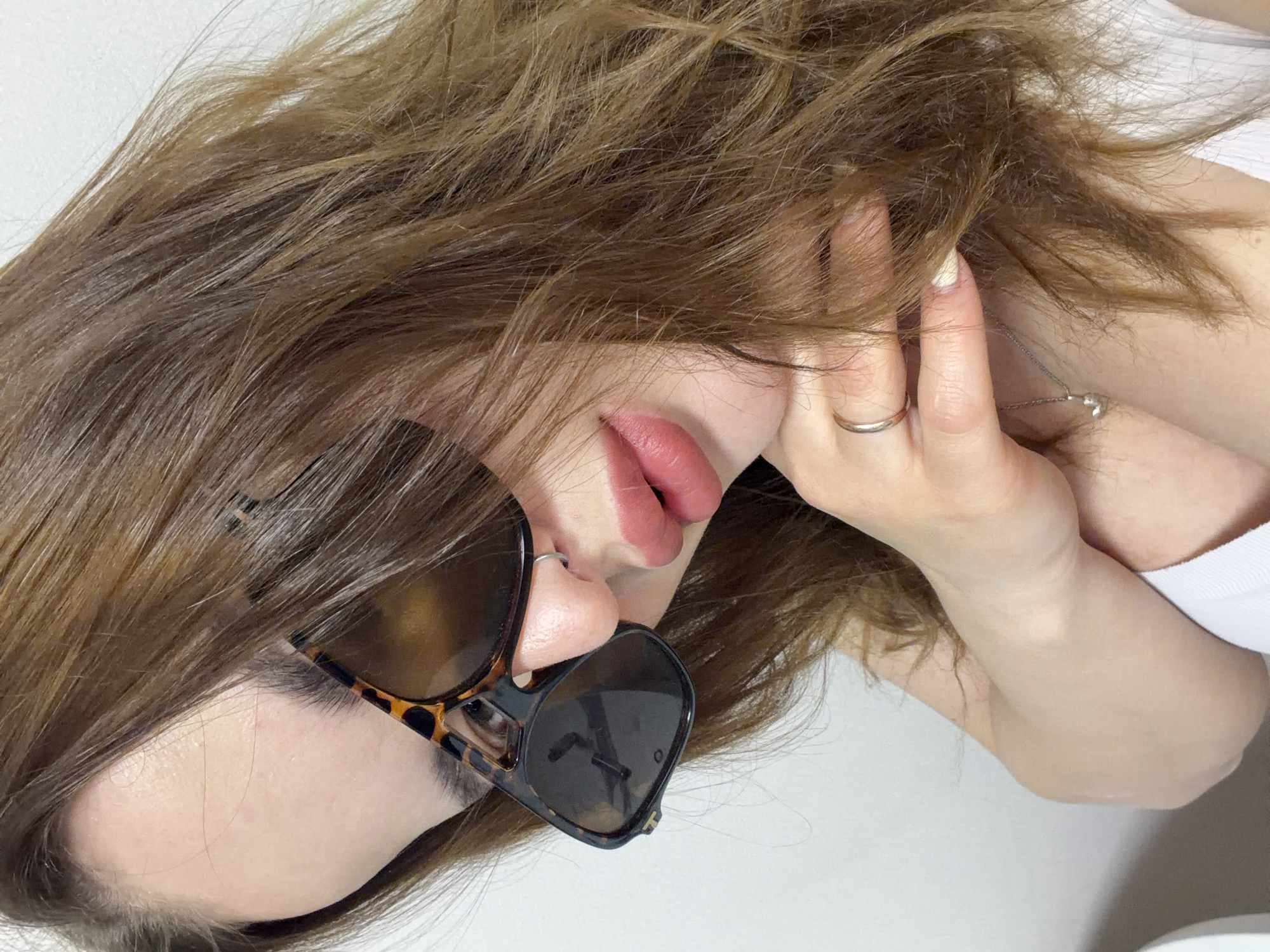 This close-up portrait features a person, likely female, against a plain white background, appearing to be lying down or leaning back with their head tilted. The subject has long, wavy, light brown hair that frames their face.
They are wearing tortoiseshell-patterned sunglasses that obscure their eyes. Their lips are painted in a reddish-pink shade and are slightly parted. A small silver nose ring is visible on the left nostril. One hand is positioned near the face, possibly touching the hair or ear, revealing a simple silver band ring on the ring finger and fingernails painted a light, neutral color. A delicate silver-colored necklace with a small pendant is partially visible near the collarbone, peeking through the hair. A section of a white top or t-shirt is visible on the upper arm and shoulder.
The lighting is soft and even, creating a calm and relaxed atmosphere. There is no visible text in the image. The setting appears to be indoors, potentially in Krasnodar, Russia.