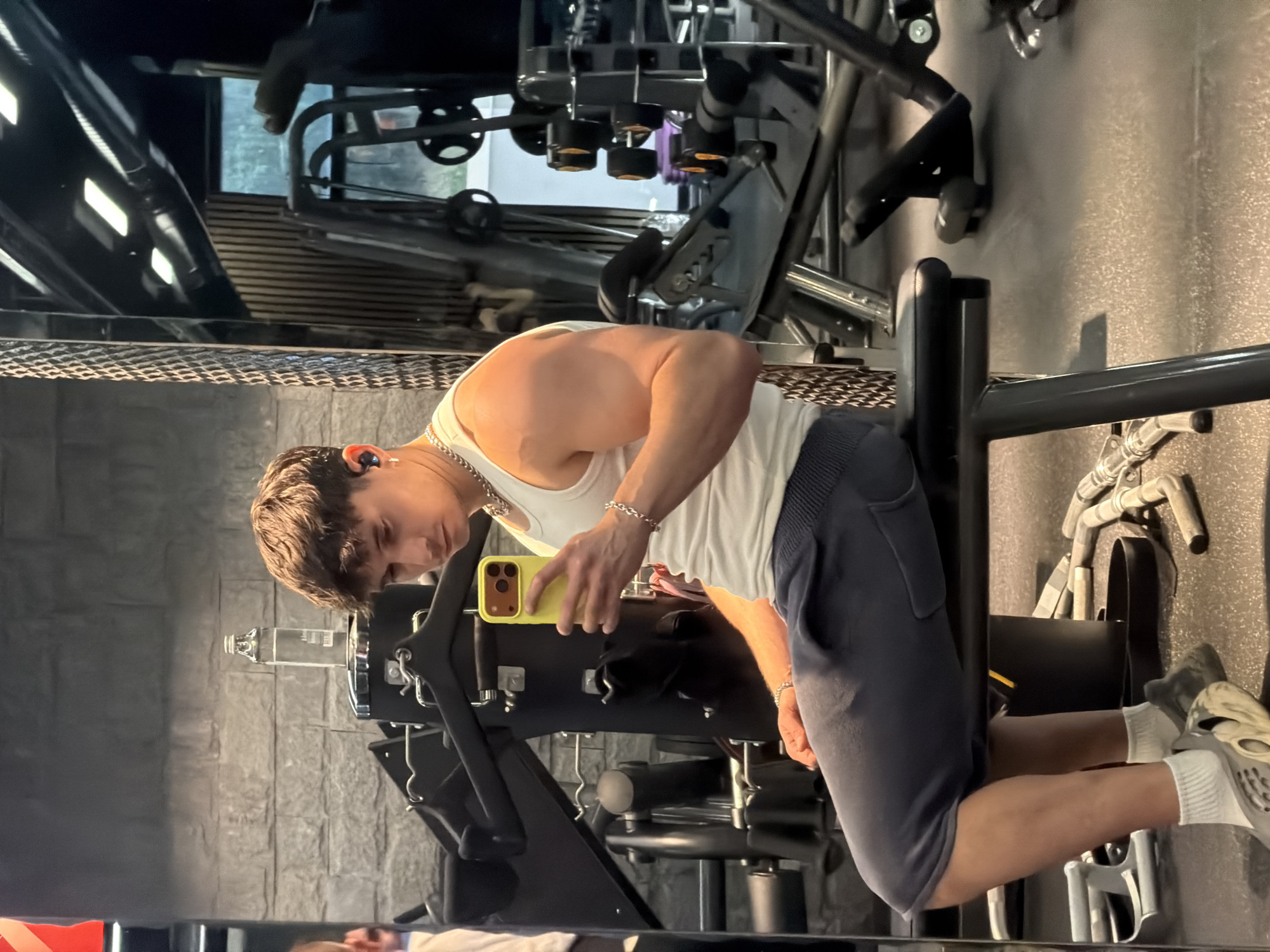 A man is in a gym setting. He is taking a photo of himself. He is wearing a white tank top, dark shorts, white socks, and sneakers. He has a chain necklace and bracelet. He is holding a yellow phone. Gym equipment is visible in the background and foreground. The location is in Sofia, Bulgaria.