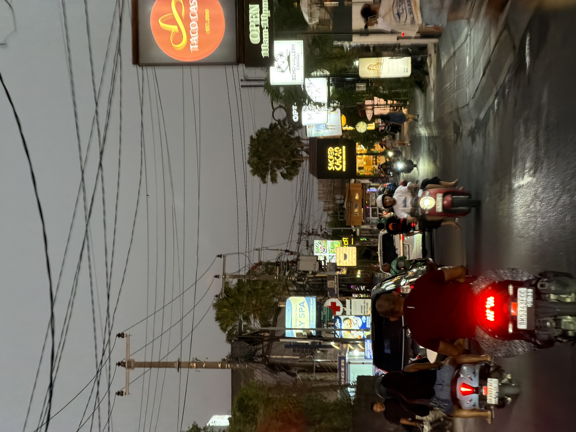The image depicts a bustling street scene in North Kuta, Bali, Indonesia, during dusk or early night under an overcast, light grey sky crisscrossed by numerous overhead utility wires and poles. The asphalt road, appearing somewhat reflective, is populated primarily by motorbikes and a few cars.
In the foreground, the rear of a red scooter (license plate DK 5343 FCC) is prominent, with its bright red taillight illuminated. A male driver, wearing a dark red shirt and patterned trousers, is seated on it. To the left, a light blue/grey scooter (license plate DK 6973 AFD) is positioned slightly ahead, ridden by a woman in a dark top and denim shorts, with bare feet. Further left, a man in a dark shirt stands near a dark-colored vehicle.
Vehicles are mostly stationary or moving slowly in a mild traffic congestion. A red scooter with a white-helmeted male driver in a white t-shirt is visible in the mid-ground, slightly to the right of the central scooter. In the background, other motorbikes and a white car are discernible amidst streetlights and illuminated signs.
The sides of the street are lined with commercial establishments featuring numerous glowing signs. On the right, a large circular orange sign for "TACO CASA EST. 2010" with a yellow sombrero icon is prominently displayed, beneath which a black sign indicates "OPEN 10am-10pm." Below these, a white rectangular sign for "ECHO GALLERY CANGGU" and another for what appears to be "MAISON NETTA" are visible. Further down the street on the right, a building with "SACRED CACAO RAW CHOCOLATE" in yellow text is illuminated. On the left side of the street, a blue sign for "Y SPA" and another with a red cross symbol are visible. Interspersed among the buildings and signs are tropical palm trees. A person in a light-colored top and denim shorts walks away from the camera on the right sidewalk, exhibiting motion blur.