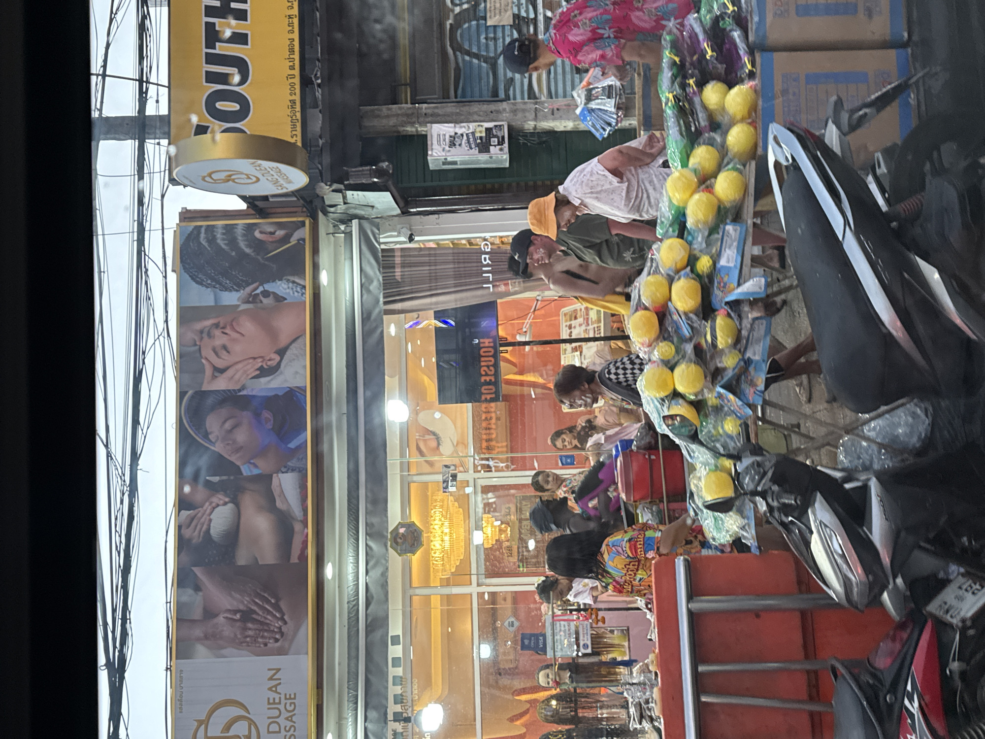 The image shows a street scene in Patong, Thailand. "Sang Duean Massage" is advertised on a sign, along with other massage businesses and shops. People are browsing a vendor selling items on the street, and motorcycles are parked nearby. A sign says "Raht Uthit 200 Year."
