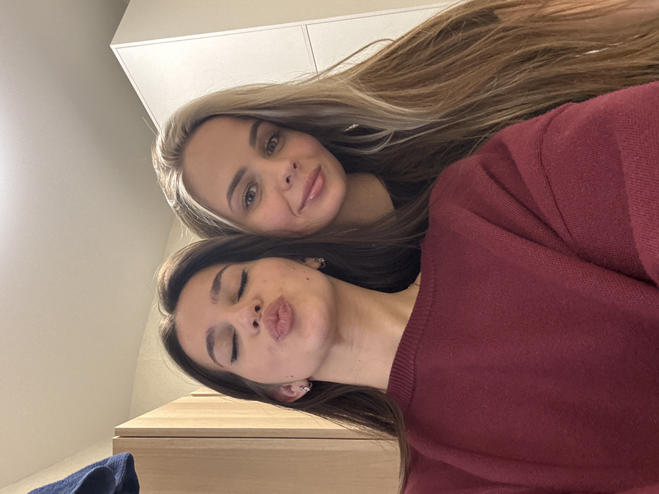 Two women are close together, posing for a selfie. The woman at the top has long, blonde hair and is smiling slightly. The woman below has dark brown hair, one eye closed, and is puckering her lips into a kiss. She wears a maroon sweater. A white cabinet is visible in the background.