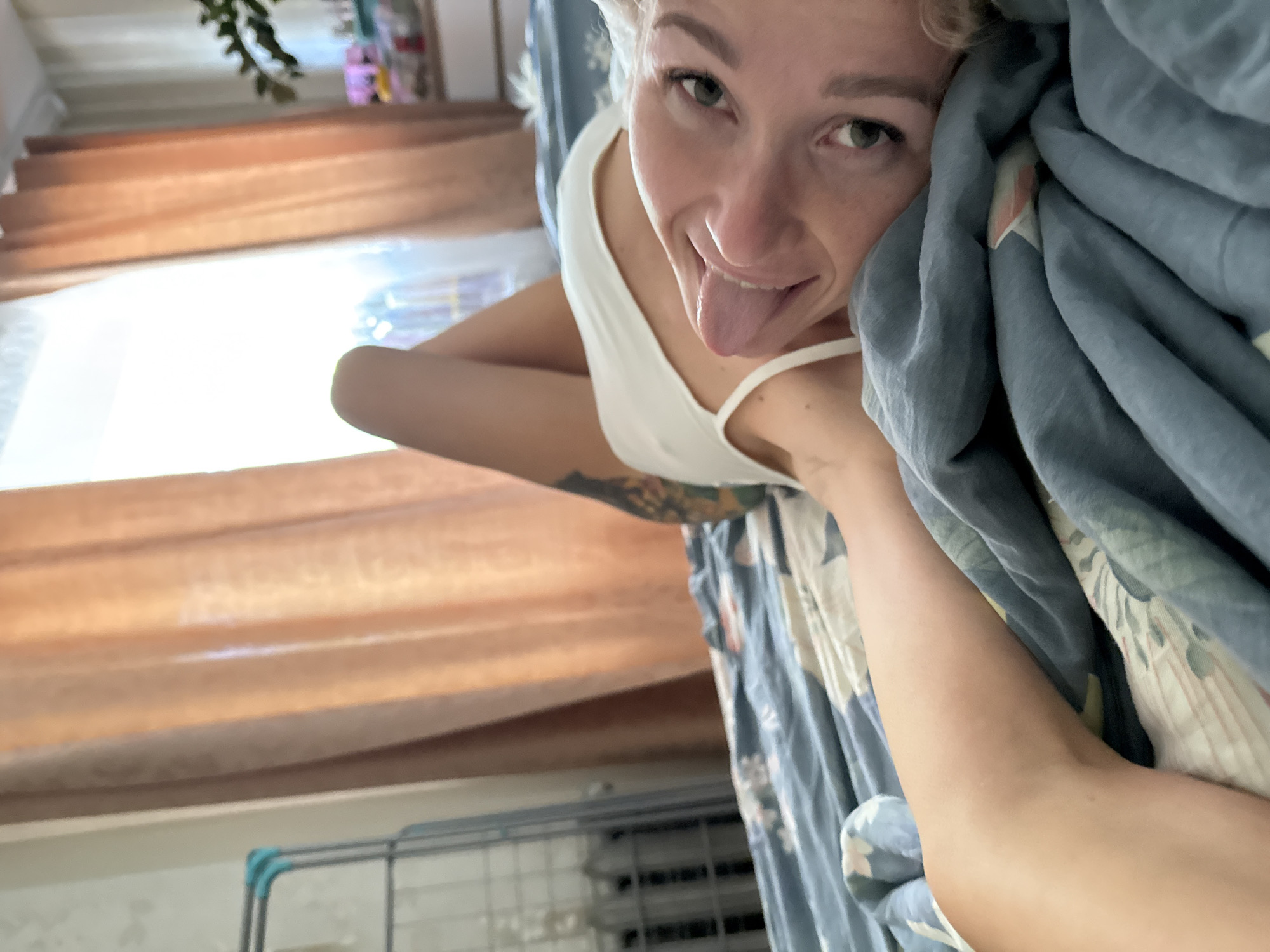 A person is lying in bed indoors, with a blue and white patterned duvet pulled up close to their face. The person is light-skinned with blonde hair and is sticking their tongue out. They are wearing a white tank top. Part of their tattooed arm is visible. In the background, there is a window with light filtering through orange curtains. Below the window, a silver drying rack is visible. The image appears to have been taken at an angle, looking down at the person in bed. The location is Chaikovsky, Russia.