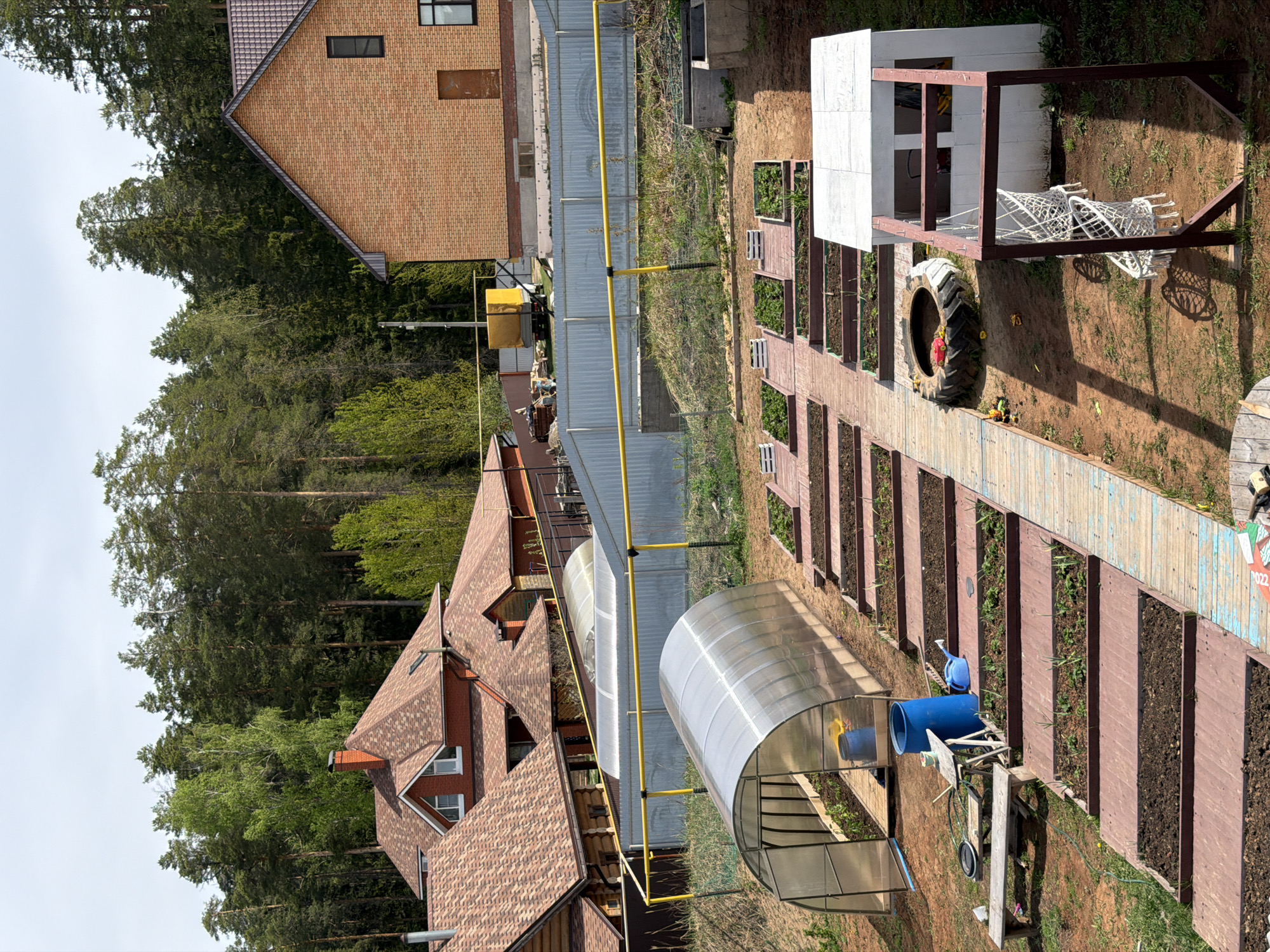 The image depicts a rural scene, likely in Chaikovsky, Russia. Two houses with red-tiled roofs are visible, surrounded by a dense forest. In the foreground, a garden features raised planting beds and a greenhouse structure. A large tire and construction materials are scattered nearby. A yellow pipeline runs through the scene. There are no people visible in the image.