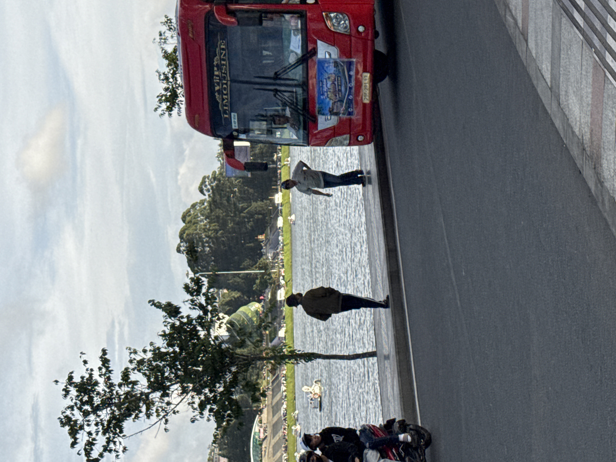 The image captures a scene along the edge of Xuan Huong Lake in Da Lat, Vietnam. A bright red VIP Limousine bus is parked on the right side of the frame, facing forward. The bus displays advertisements on its windshield and front. To the left of the bus, two men are standing by the water's edge. One man, wearing a light-colored shirt and dark pants, is walking away from the viewer. Another man, in a dark jacket and pants, is facing the lake with his hands behind his back. In the foreground, a motorcycle with two passengers is parked, with one passenger sitting on the back and the other in front. The lake stretches across the mid-ground, with several small white boats visible on its surface. In the background, a line of trees and buildings is visible, with a cloudy sky overhead. The overall scene suggests a casual, everyday moment in a popular tourist area.