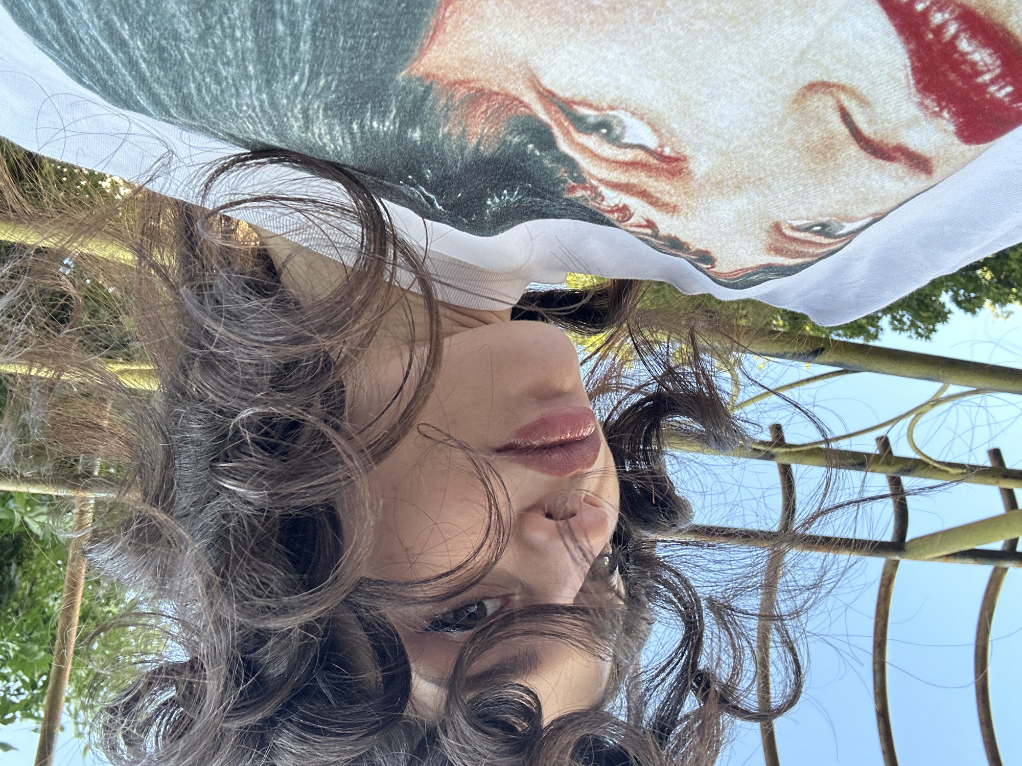 A young woman with curly dark brown hair is looking down at the camera, her face tilted. She is wearing a white t-shirt with a printed image of a woman's face with red lipstick. Her hair is partially obscuring her face and is styled in loose curls. She is outdoors, with a bright blue sky and some greenery visible in the background. The image appears to be taken from a low angle, looking upwards. There is a metal structure with a curved design behind her, which may be part of a garden trellis or similar outdoor decoration. The lighting suggests it is daytime. The specific location in Sofia, Bulgaria cannot be determined from the image.