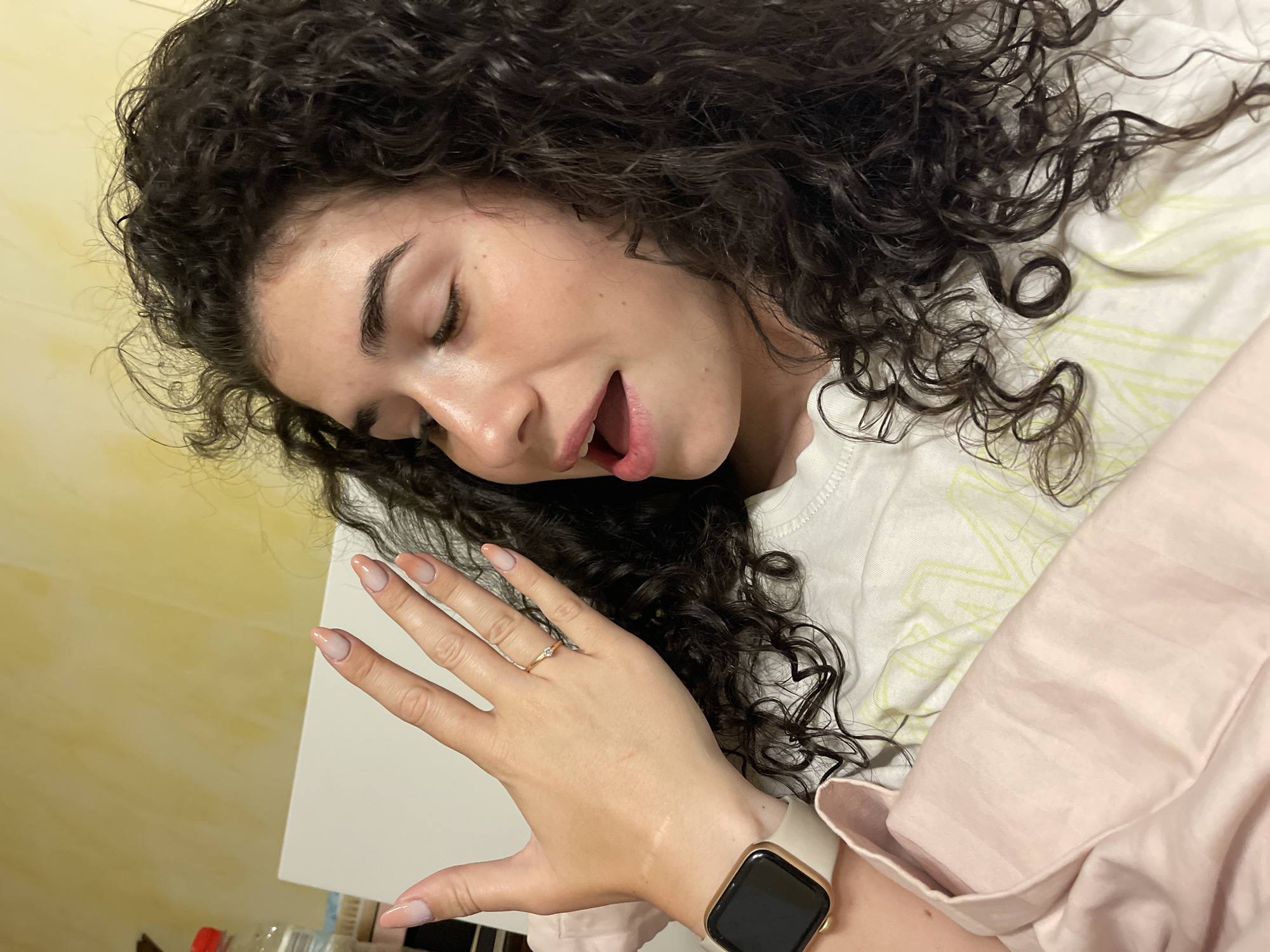 A young woman with dark, curly hair is lying in bed, looking to the side with her mouth slightly open. She is wearing a white t-shirt with a yellow graphic design and has a pink blanket over her. Her right hand is raised, displaying her fingernails which are painted with a nude to pink gradient. She is also wearing a gold ring with a small diamond on her left ring finger and a smartwatch on her left wrist. The background appears to be a yellow wall. The scene is indoors, in a bedroom. There is no specific indication of the location being Sofia, Bulgaria.